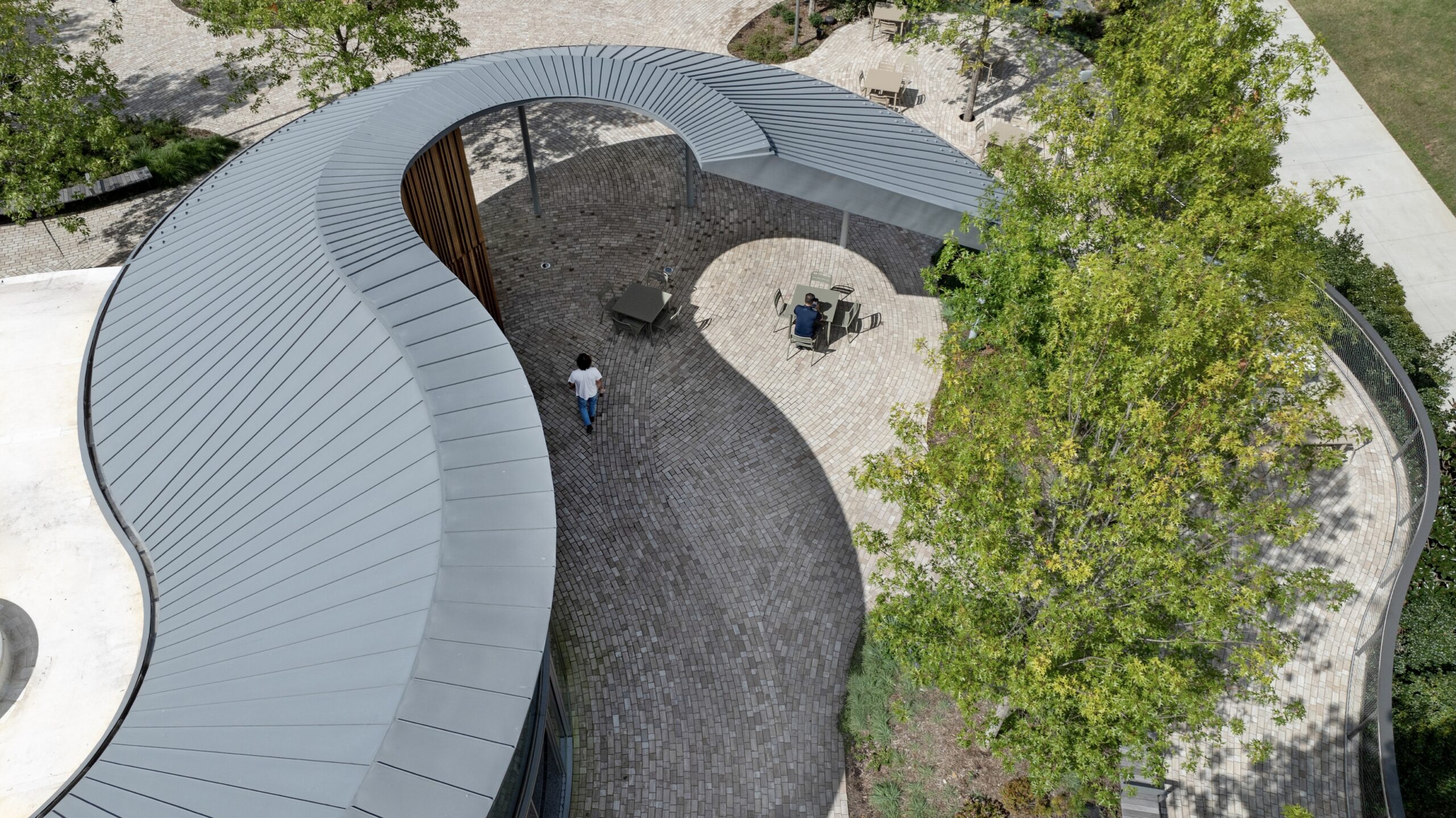 Aerial view of a curved building with a metal roof at a Camp for the Blind, featuring a brick patio, two people walking, and trees providing shade.