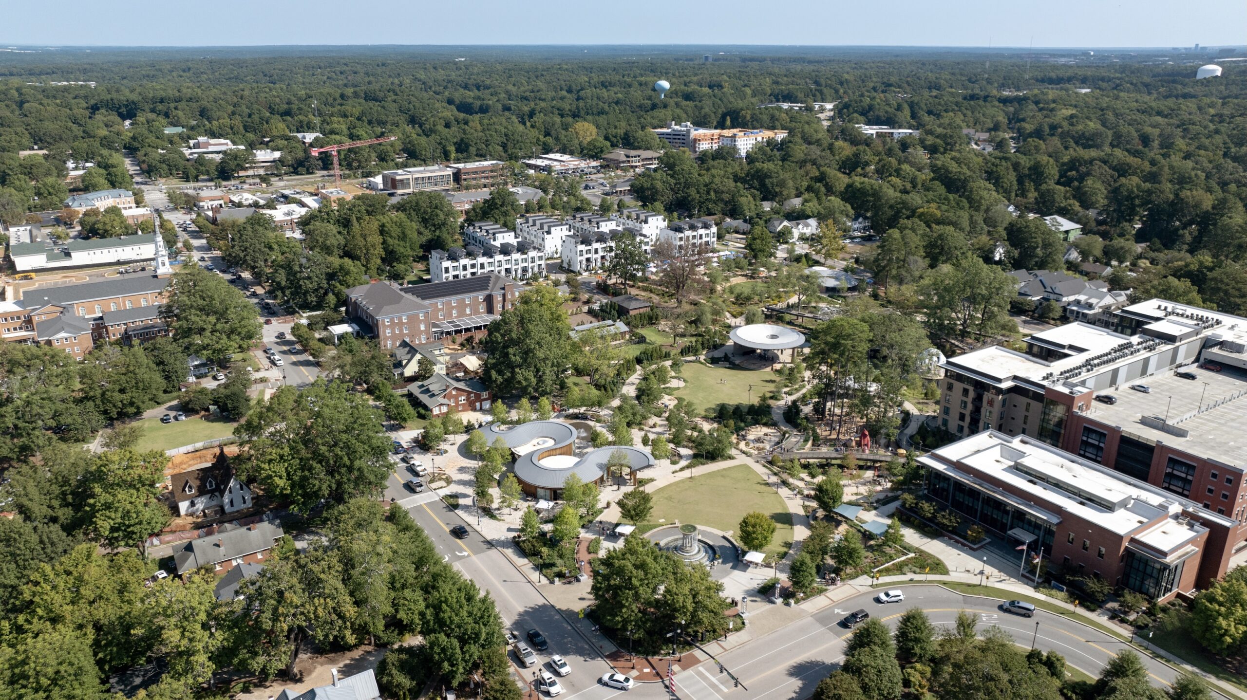 Aerial view of a suburban downtown area with tree-lined streets, commercial buildings, a park with winding paths near a renowned Camp for the Blind, and parking lots.