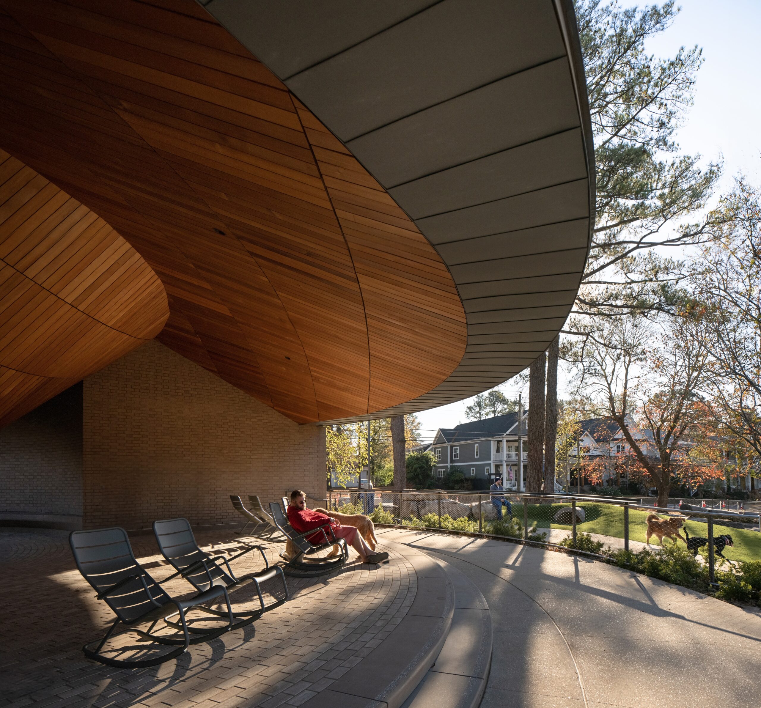 A person sits on a lounge chair under a large modern overhang with wooden ceiling panels, looking out at a landscaped area with trees and nearby houses—an inviting, tranquil scene reminiscent of the peaceful atmosphere found at a Camp for the Blind.