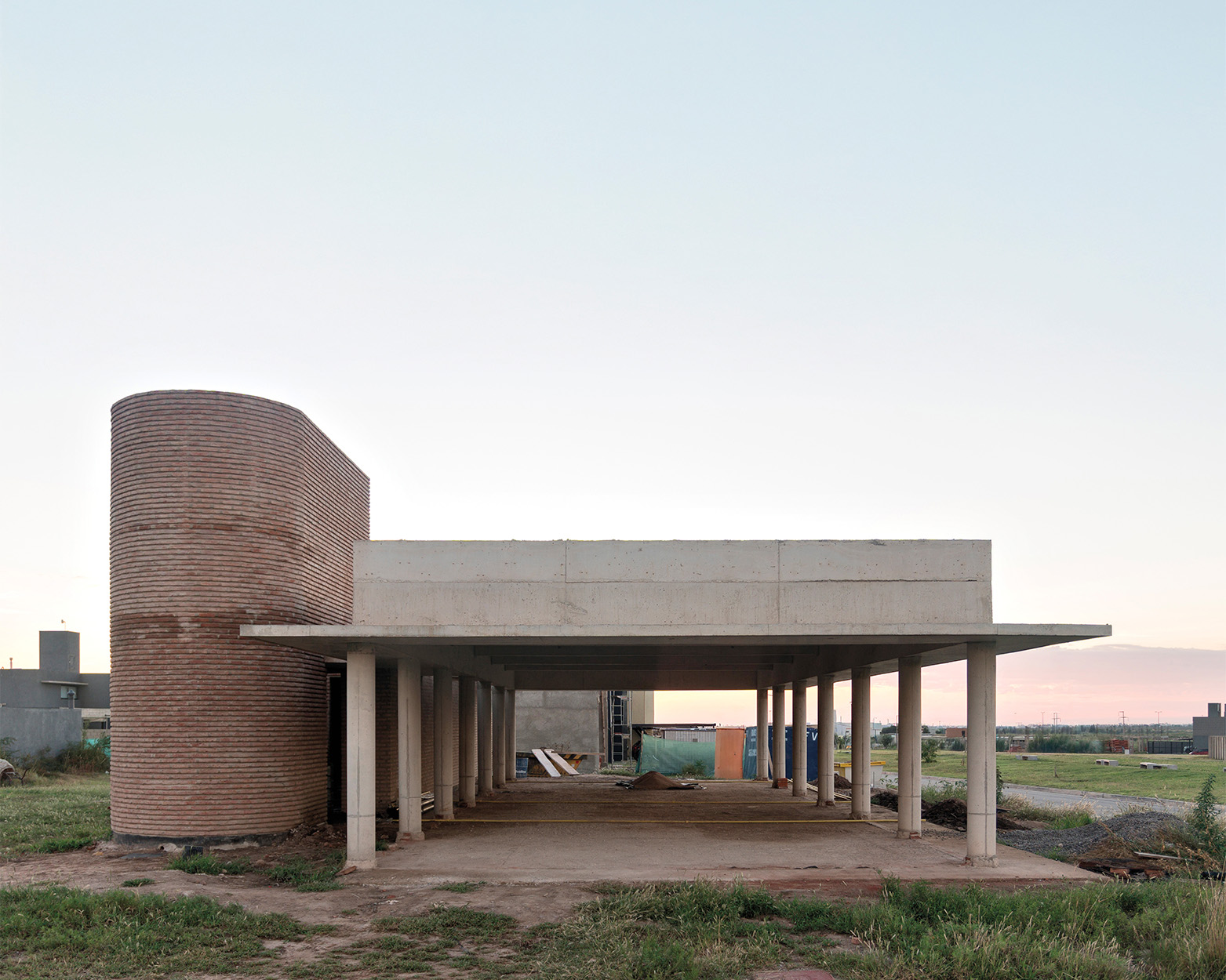 Single-story modern building with a flat roof supported by columns and a cylindrical brick structure on the left, showcasing Latin Matter influences, set in a grassy area under a clear sky.