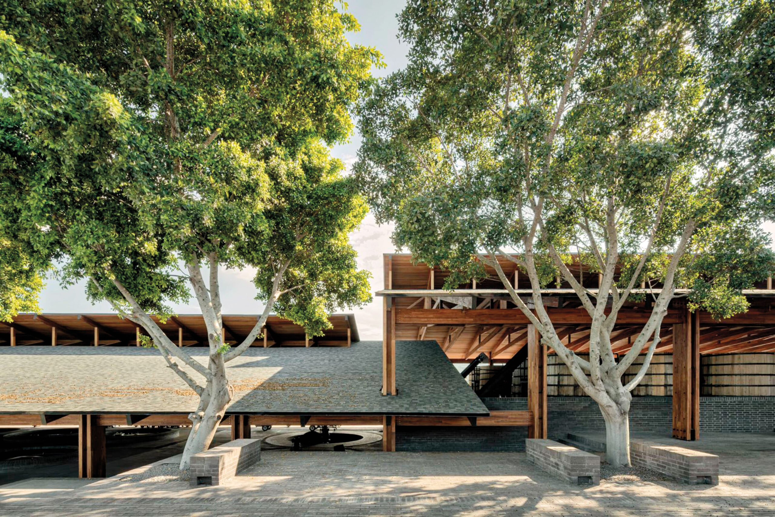 Modern building with sloped shingle roofs and open wooden structures, designed by Neri&Hu, surrounded by large trees and brick pavement.