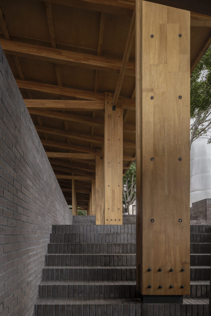 A staircase with dark brick steps passes under a wooden roof, supported by large vertical wooden beams in the signature style of Neri&Hu; trees are visible through openings on the right.