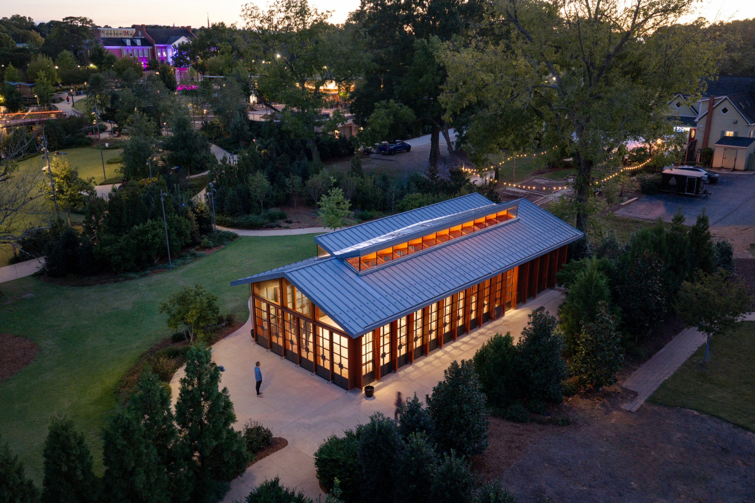 A modern glass and wood building lit from within sits among trees and pathways at dusk at a Camp for the Blind, with a few people walking nearby.