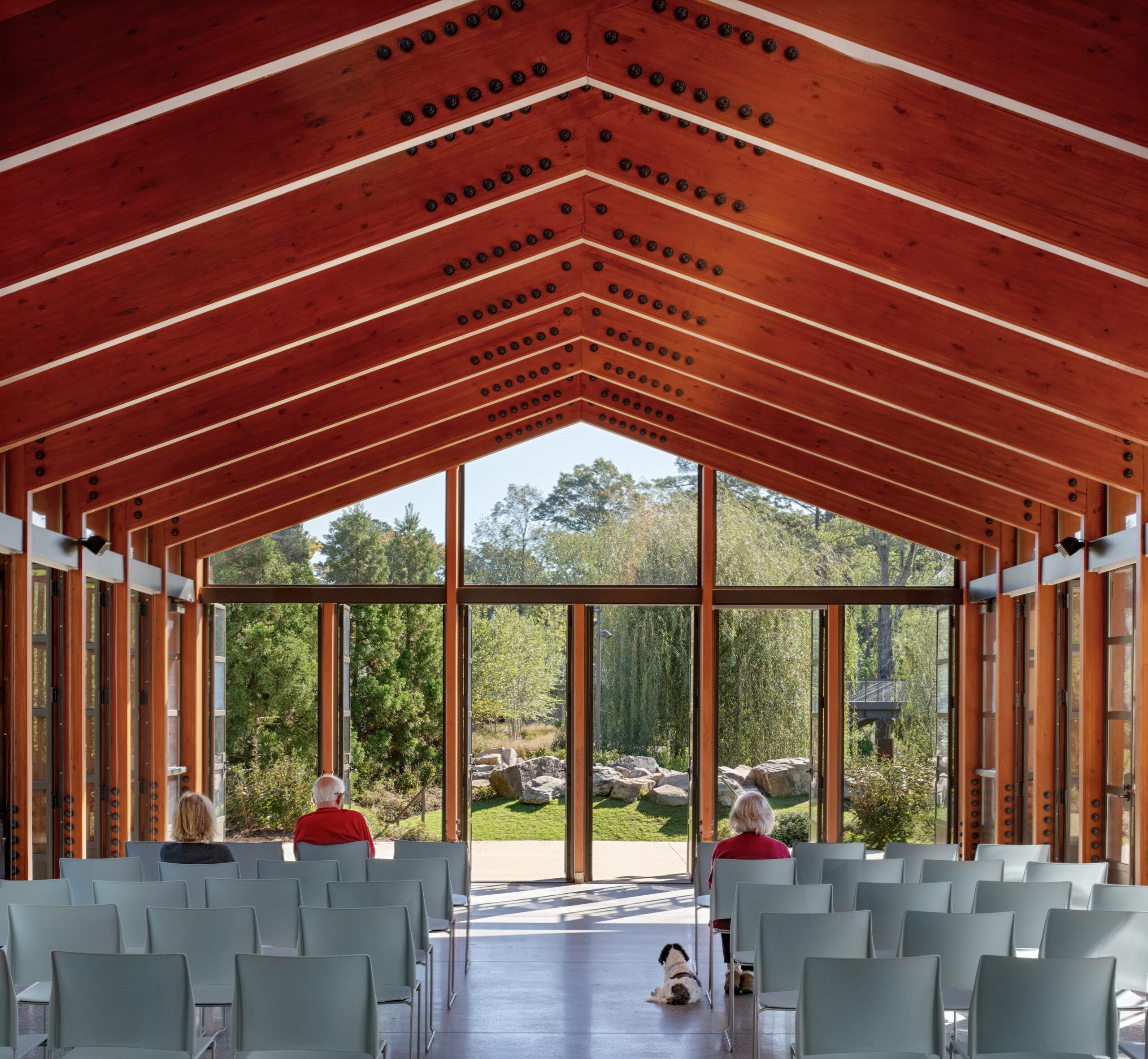 Three people and a dog sit facing large glass windows in a modern wooden chapel at a Camp for the Blind, with rows of empty blue chairs and garden views outside.