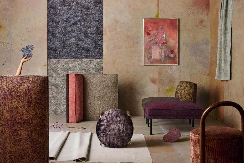 Modern living room with abstract patterned furniture, textured rugs, a wall painting, and a hand reaching in from the left holding a blue object. Walls and decor feature muted earth tones.