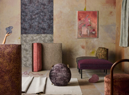 Modern living room with abstract patterned furniture, textured rugs, a wall painting, and a hand reaching in from the left holding a blue object. Walls and decor feature muted earth tones.