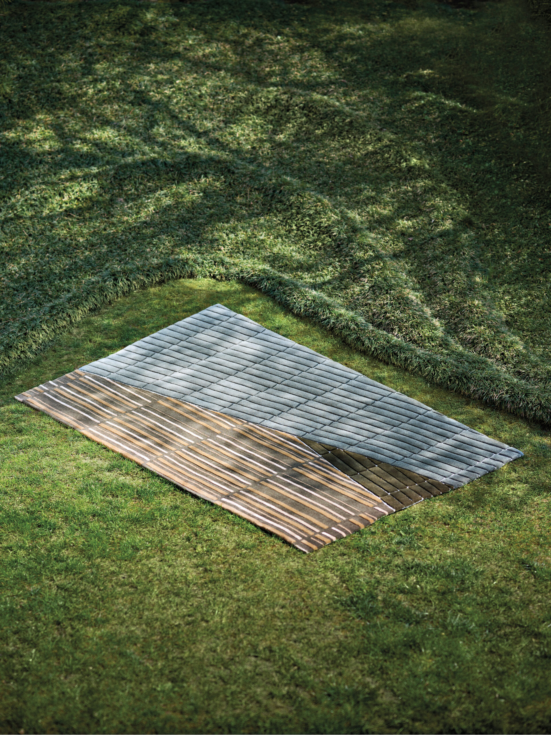 A rectangular section of grass features an artistic installation resembling a partially peeled surface, exposing a pattern of gray and brown slats beneath. Product collection by Jaipur Rugs.