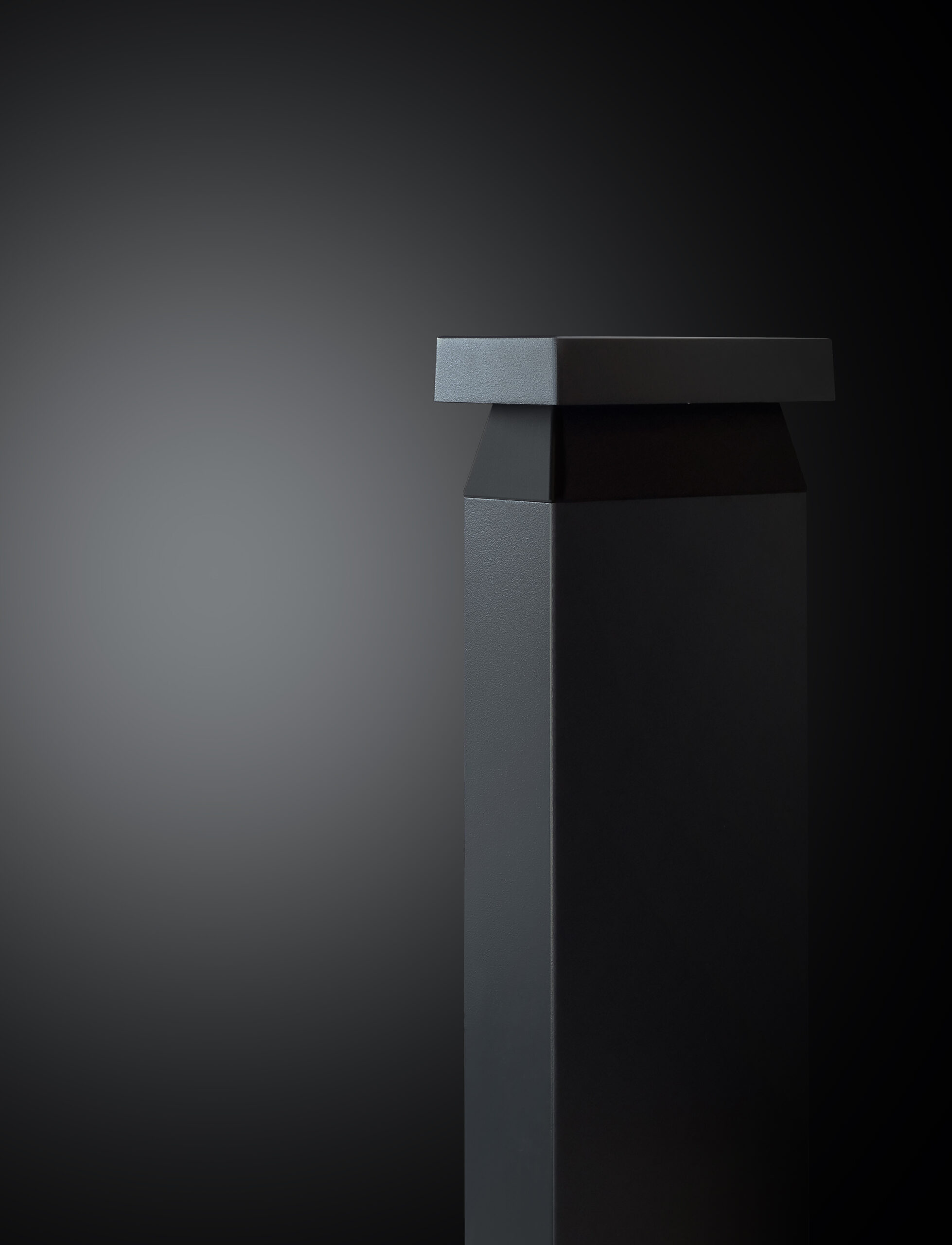 Neuro-Inclusive Lighting: A minimalist, matte black pedestal with sharp edges stands against a gradient dark background, positioned slightly off-center to the left.