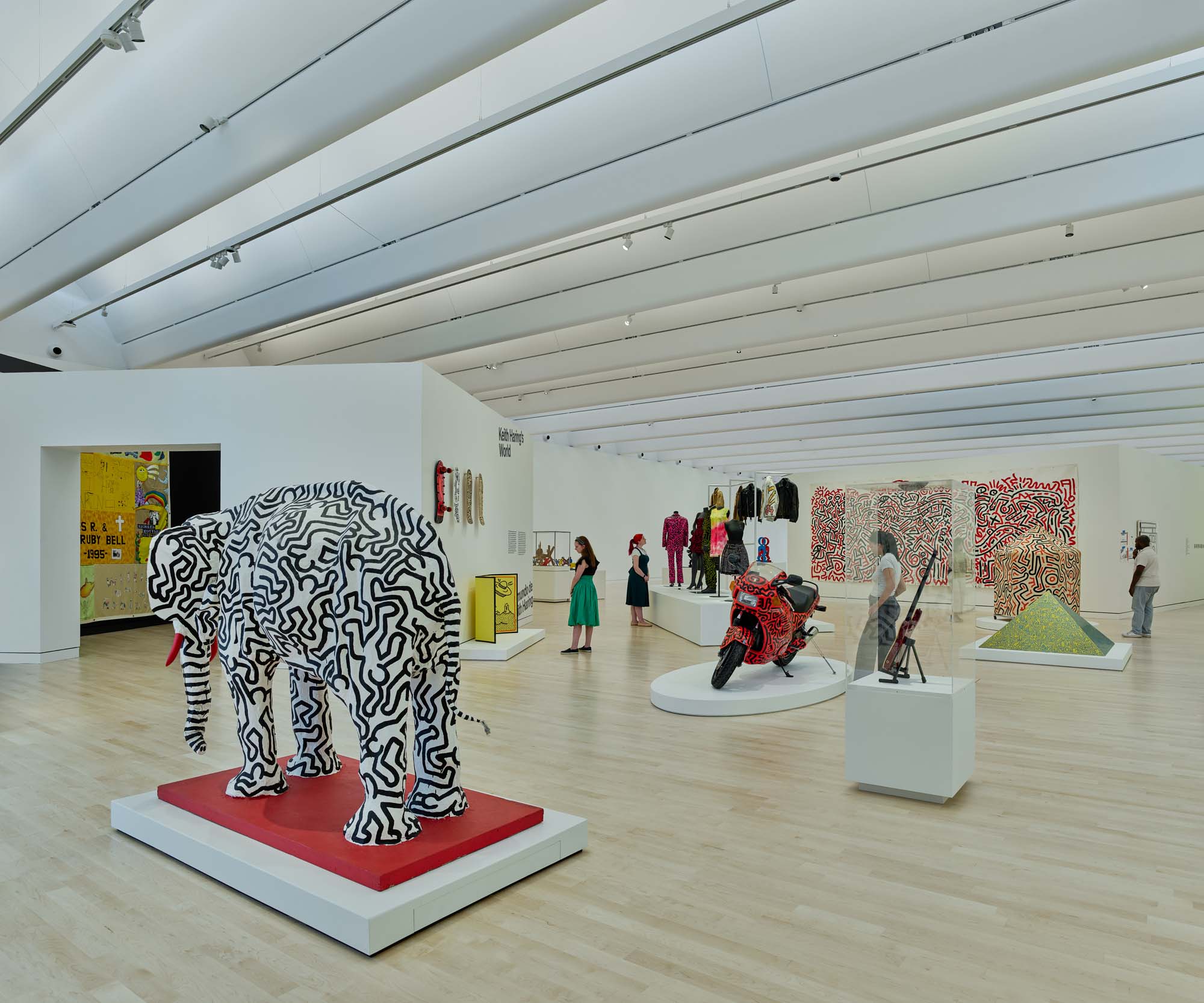 Modern art gallery with a patterned elephant sculpture, colorful motorcycle, various art pieces, and visitors viewing the exhibits in a bright, spacious room.
