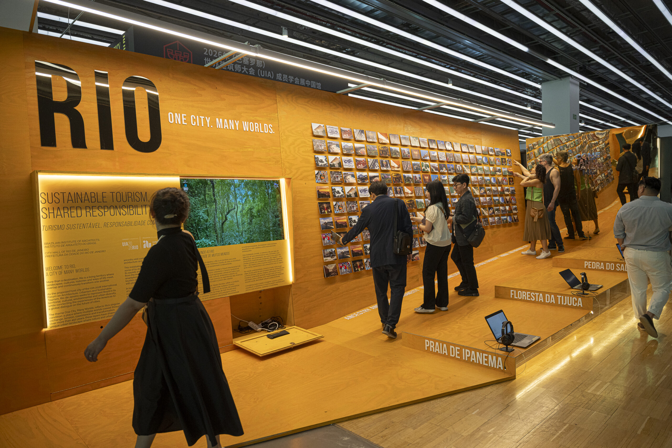 Exhibit with visitors displays "RIO: One City, Many Worlds," featuring photos, text panels, and a screen about sustainable tourism in Rio de Janeiro on bright orange walls at 3daysofdesign.