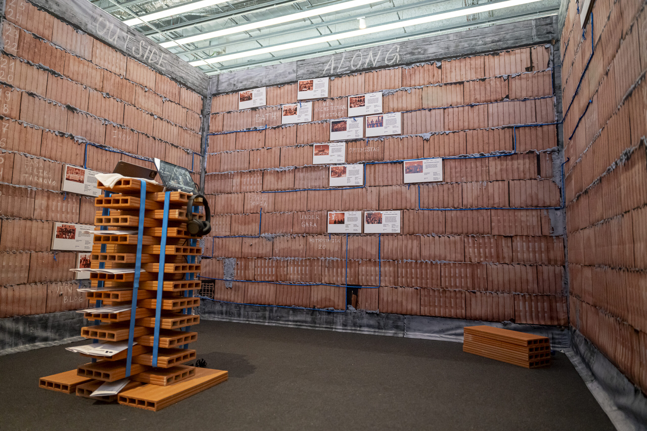 A room with red brick walls, printed papers on the back wall, and a stacked pile of bricks with blue straps on the floor sets the stage for 3daysofdesign. Fluorescent lights glow overhead.