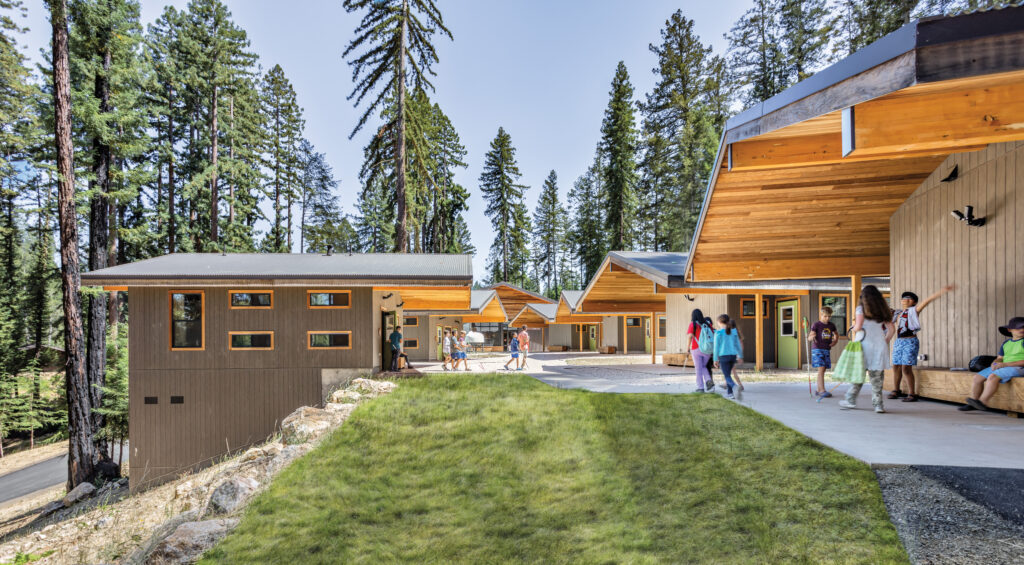 Camp for the Blind: People walk and gather near modern wooden cabins surrounded by tall pine trees on a sunny day.