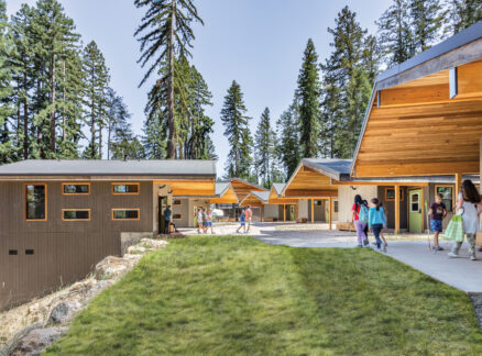 Camp for the Blind: People walk and gather near modern wooden cabins surrounded by tall pine trees on a sunny day.