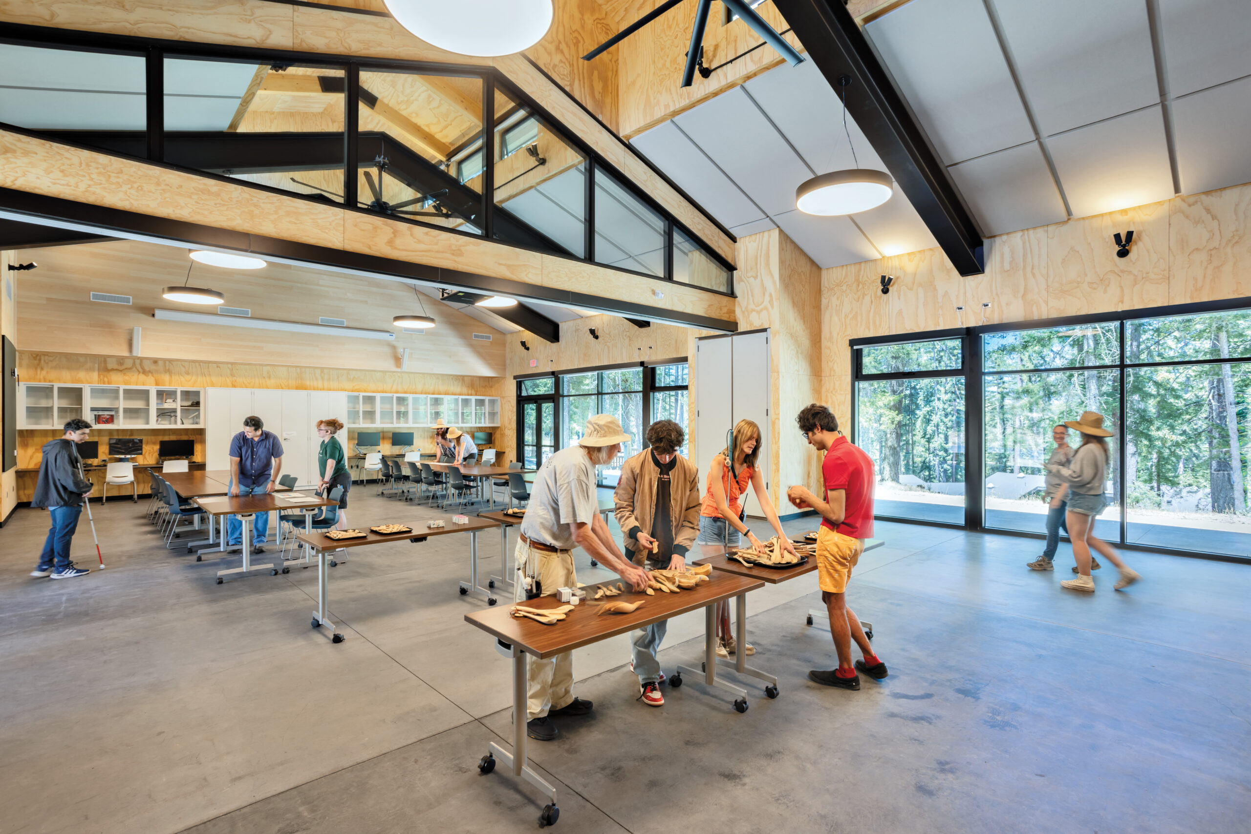 People work on projects at tables in a spacious, modern camp for the blind classroom with large windows, wood-paneled walls, and views of trees outside.