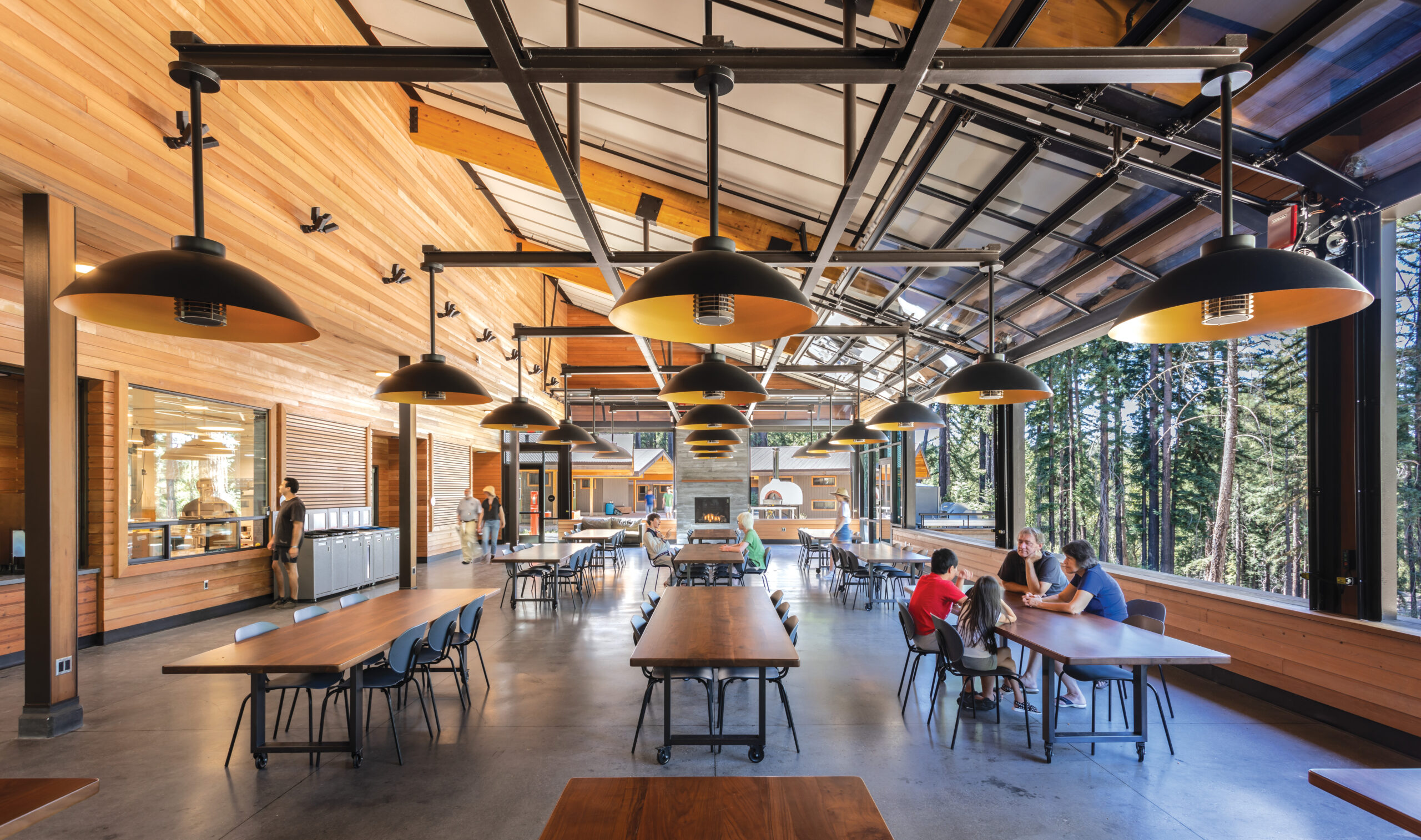 Spacious cafeteria with high ceilings, large windows, wooden walls, and several people seated at tables in a bright, open setting.