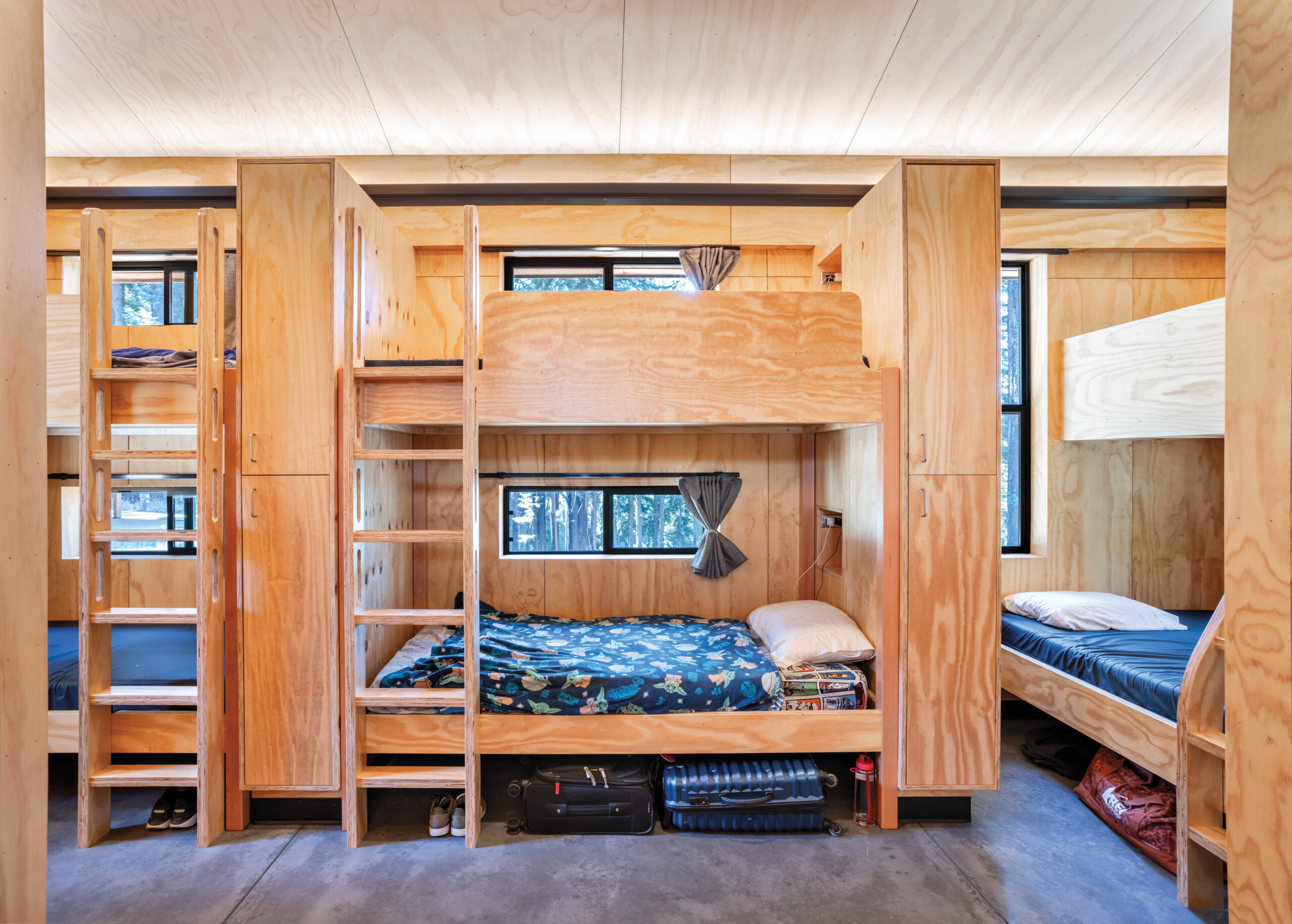 Wooden bunk beds with bedding line a cabin room, each with a window, ladder, and storage space below; suitcases and personal items are visible under some beds.