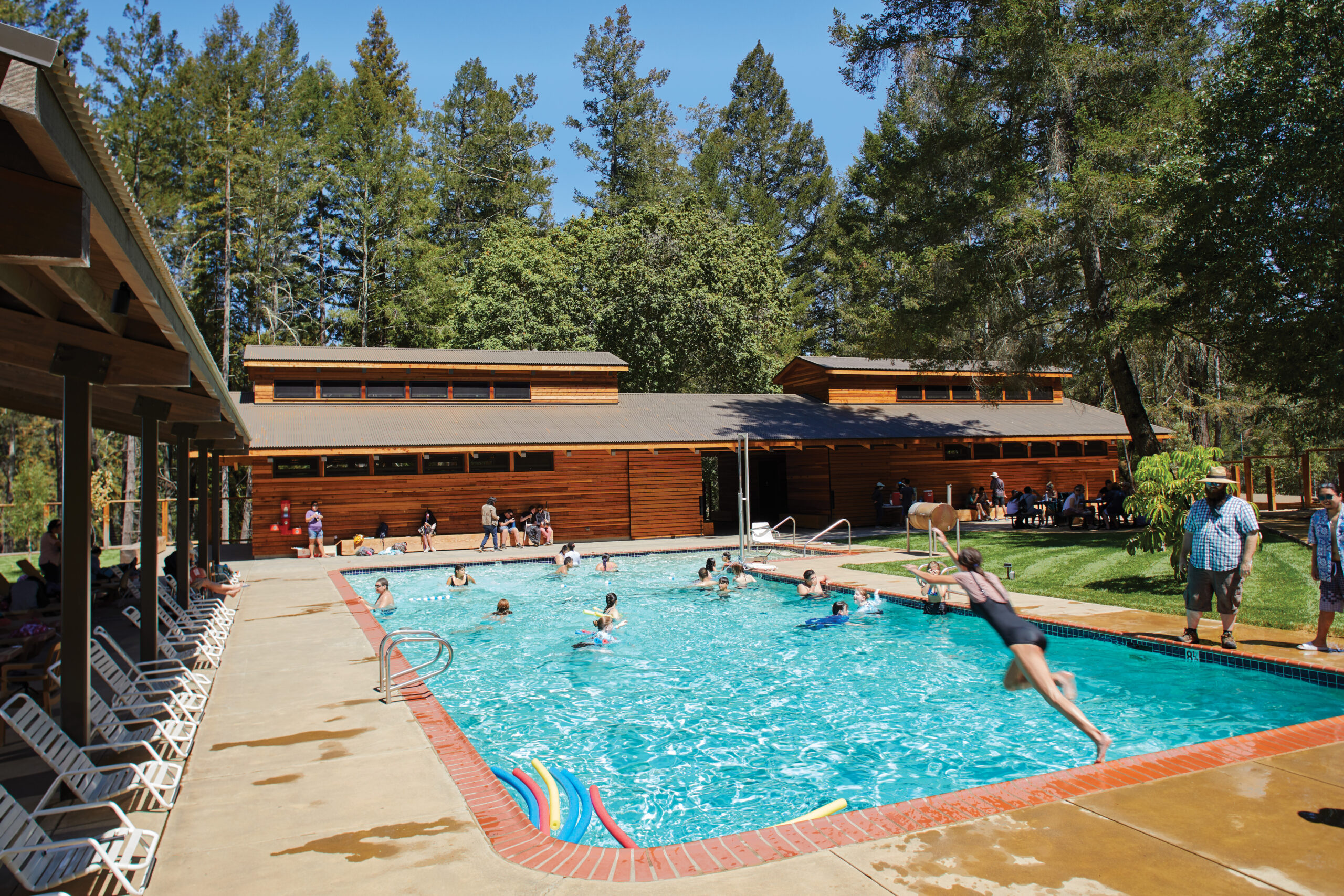 People swim and play in an outdoor pool near a wooden camp for the blind building surrounded by trees; one person is jumping into the pool.