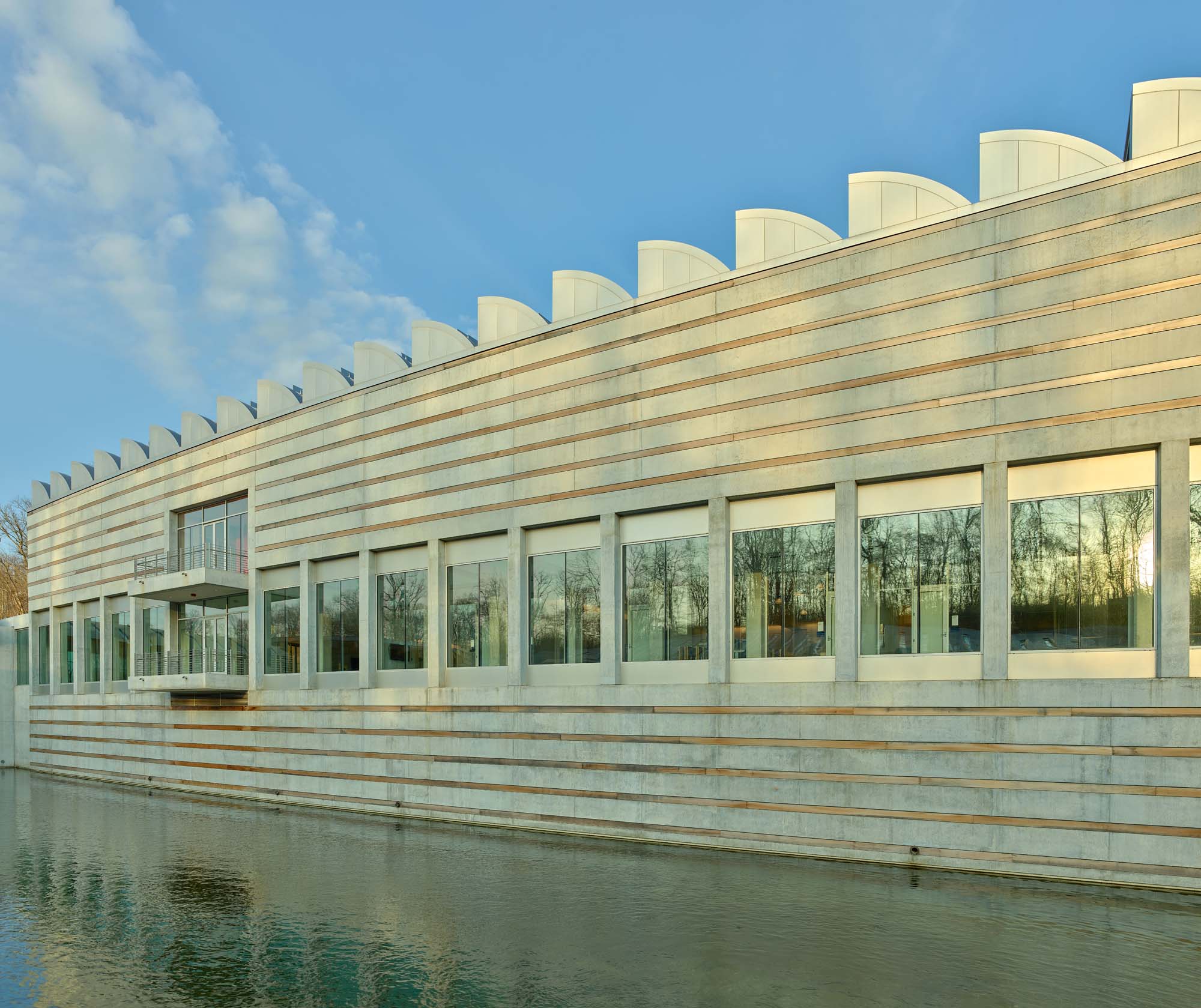 Modern building with large windows and a rippled roofline, reflected in a body of water under a clear blue sky.