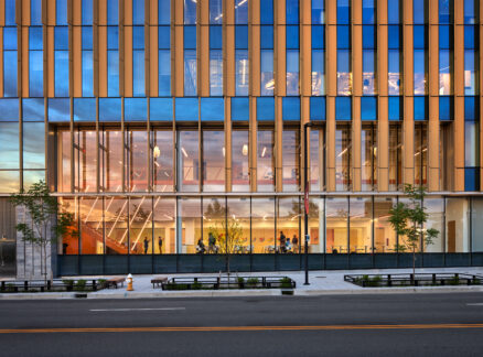 A modern building with large glass windows and vertical gold-colored fins, showing a lit interior with visible people and staircases at dusk.