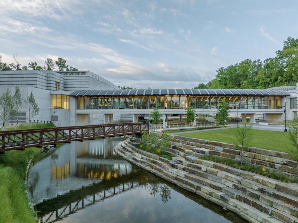 Modern building with glass and concrete facade, surrounded by trees, sits beside a creek with a footbridge and tiered stone landscaping in the foreground.