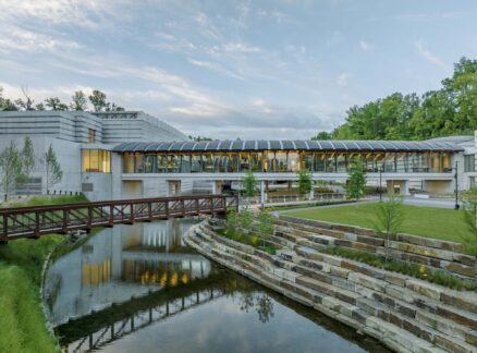Modern building with glass and concrete facade, surrounded by trees, sits beside a creek with a footbridge and tiered stone landscaping in the foreground.