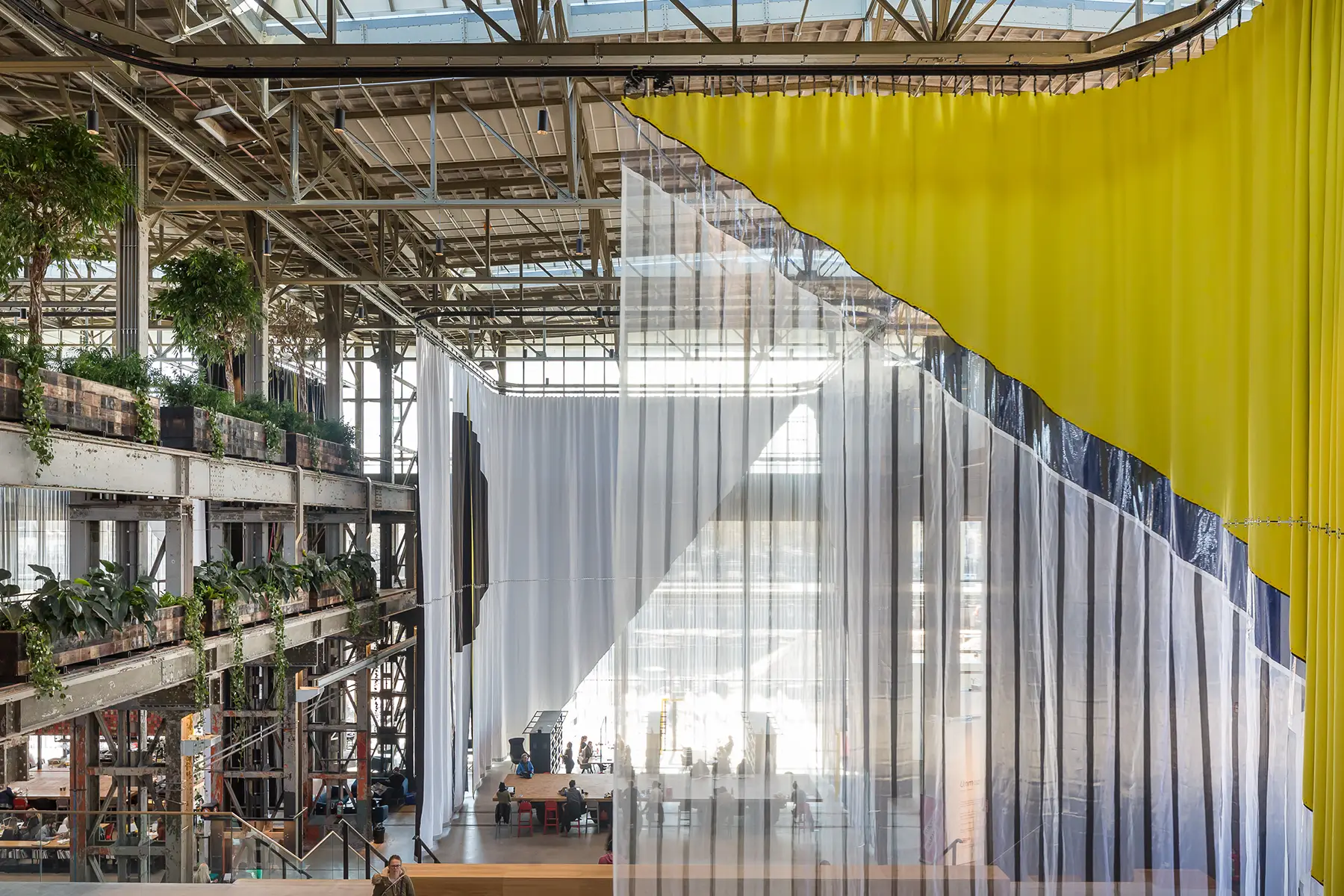Large industrial interior with high ceilings, hanging plants on metal shelving, and translucent curtains in white, black, and yellow geometric patterns dividing the space—perfect for a 3daysofdesign-inspired setting.