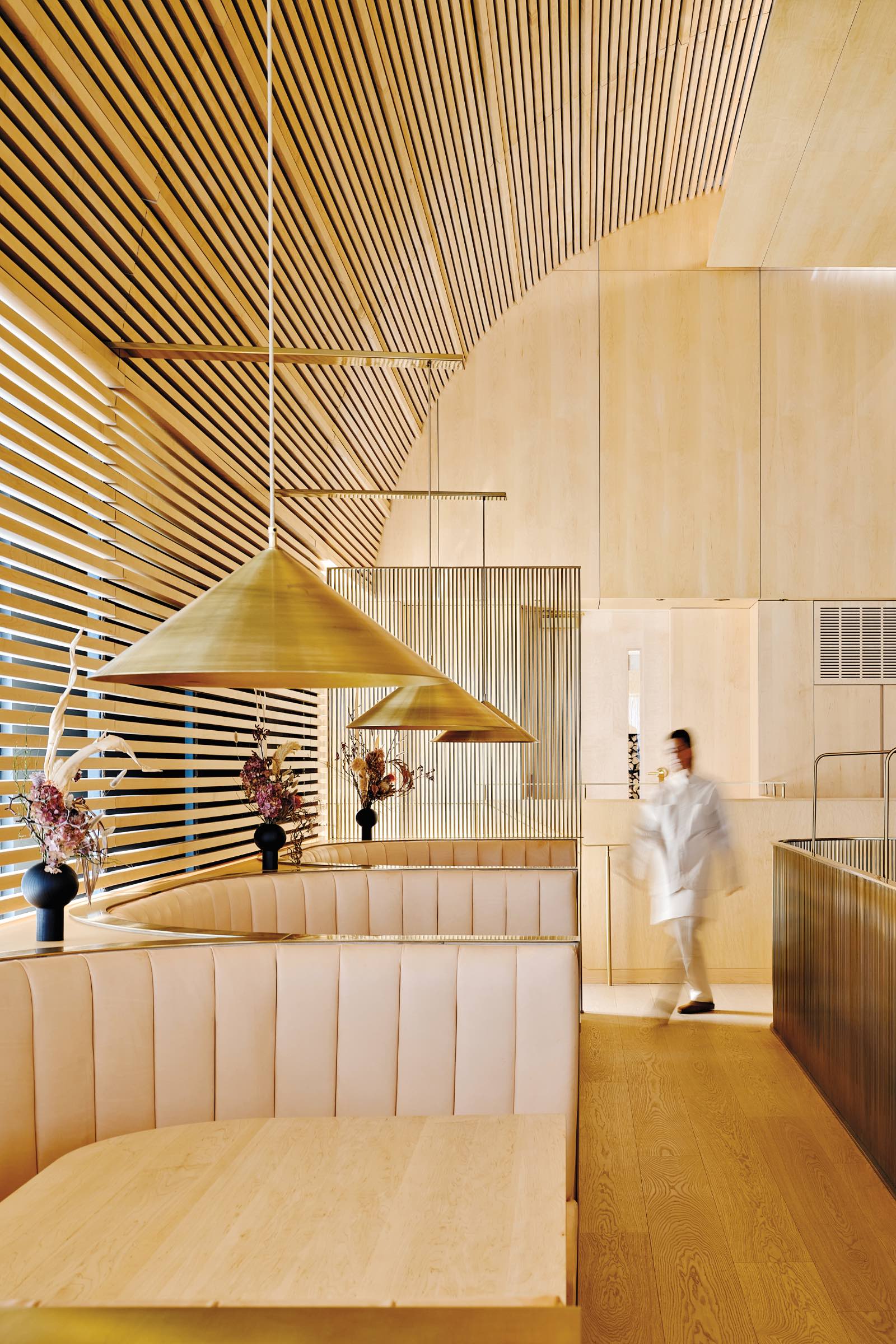 Modern restaurant interior by Omar Gandhi with light wood paneling, beige booth seating, gold pendant lights, and a person in white walking past minimalist floral arrangements.