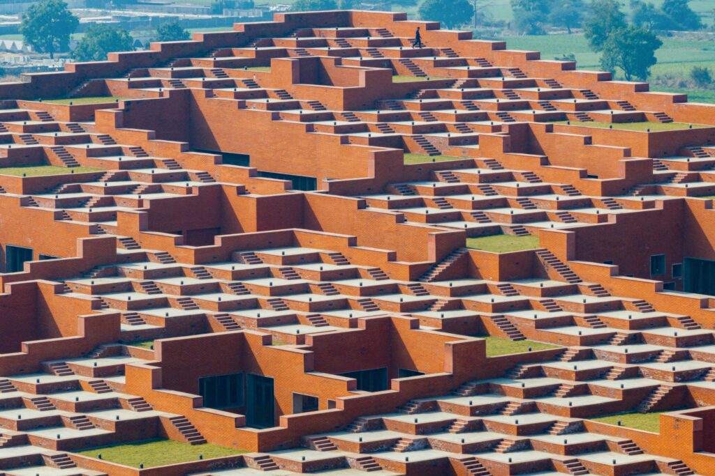 A large, terraced brick building with repeating geometric patterns and staircases. One person stands on the rooftop. Green fields are visible in the background.