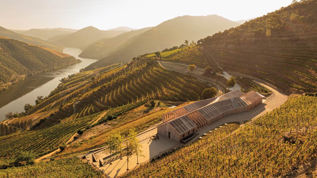 A modern building sits among terraced vineyards on rolling hills beside a winding river under a hazy sky.