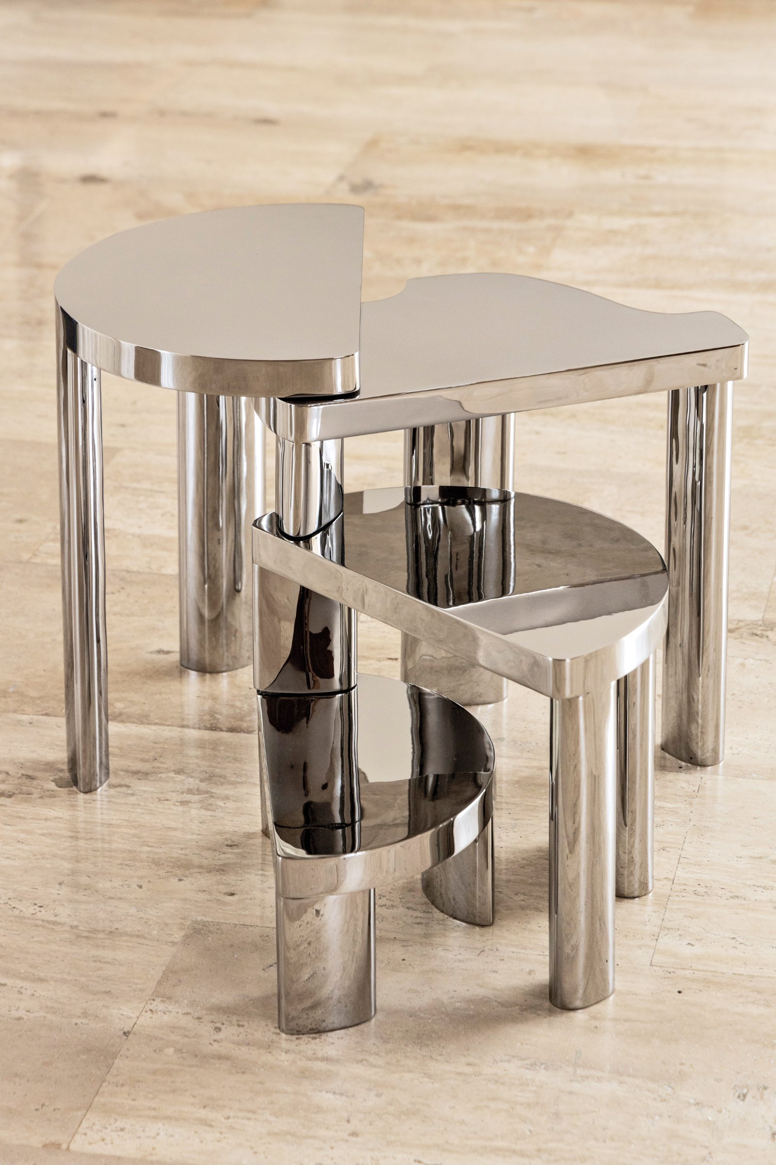 Two metallic nesting tables with asymmetrical tops are stacked on a light-colored marble floor. The tables have shiny surfaces and cylindrical legs.