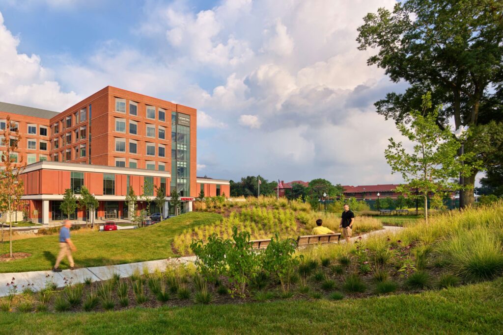 A modern brick building with large windows overlooks a landscaped lawn with benches, trees, and walking paths under a partly cloudy sky. Three people are visible in the scene.