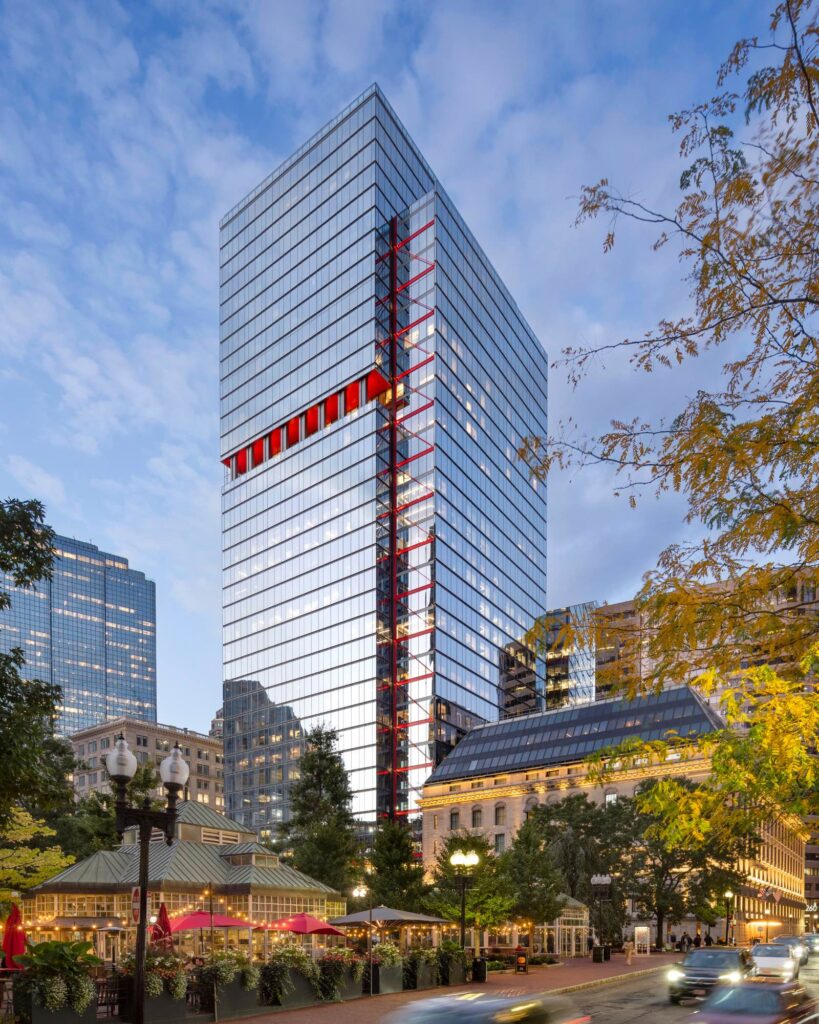 A modern glass office tower with a red structural accent on its facade stands among older buildings and trees under a partly cloudy sky.