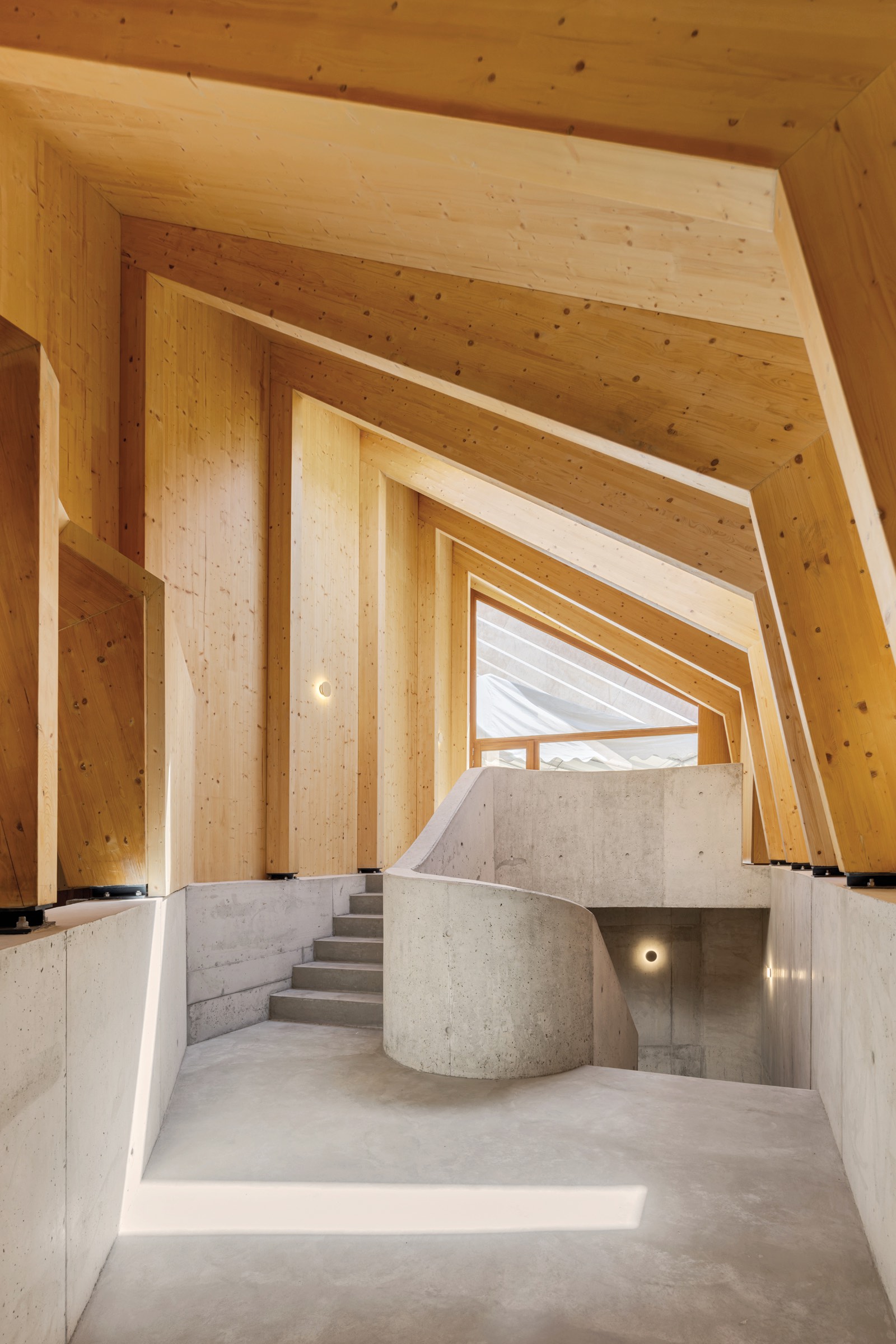 Atelier Sérgio Rebelo: Interior view of a modern building with geometric wooden ceiling panels, concrete floors, and a curved staircase leading to an upper level with large windows.