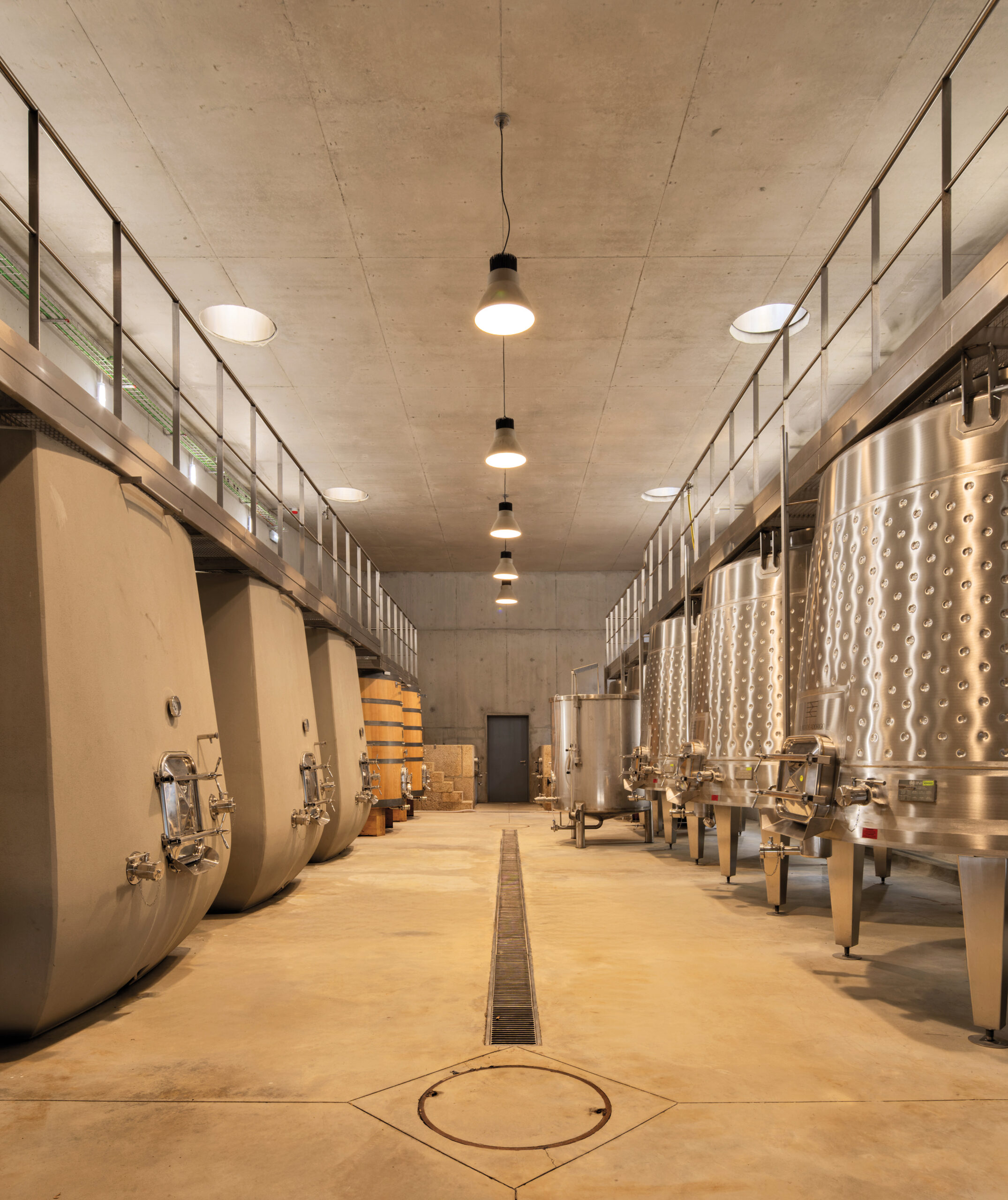 Interior of a modern winery by Atelier Sérgio Rebelo with large metal and concrete fermentation tanks lining both sides of a well-lit, clean corridor.