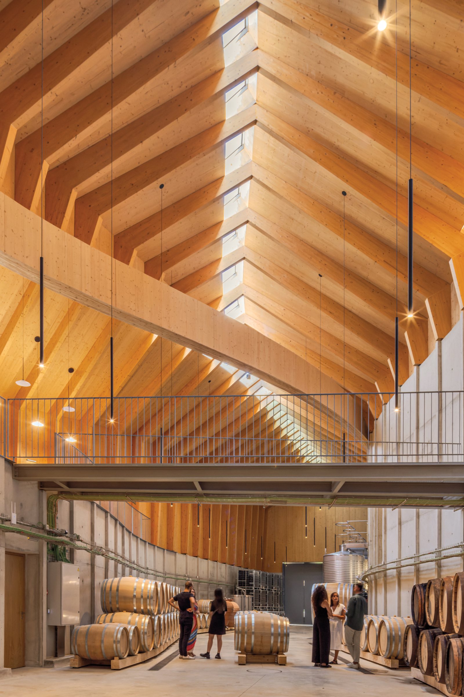 Spacious modern winery interior by Atelier Sérgio Rebelo with wooden ceiling, skylights, wine barrels, and several people standing and talking on the main floor.