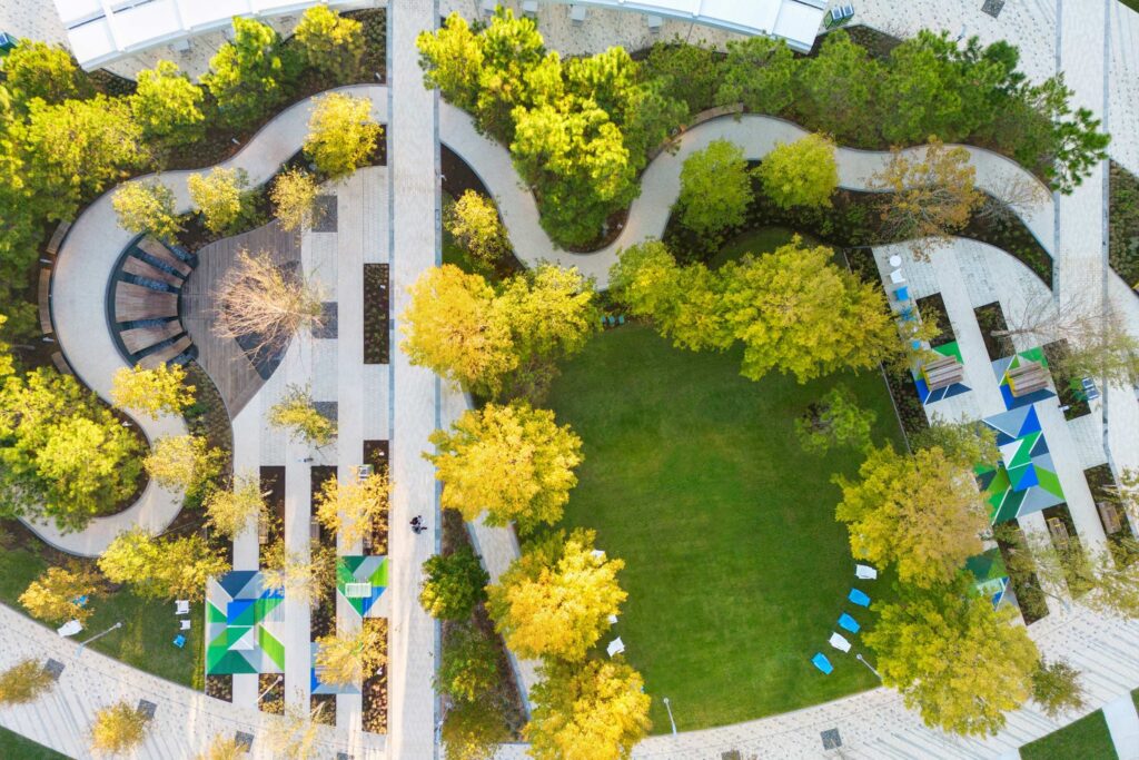 Aerial view of a landscaped park with winding paths, green lawns, trees, shaded seating areas, and geometric-patterned umbrellas.