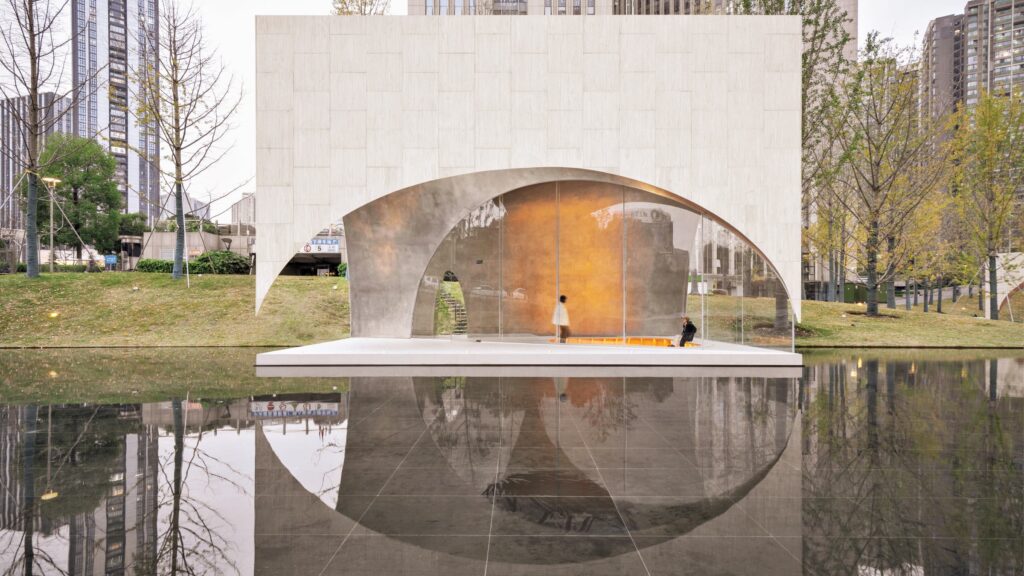 A modern structure with arched openings and textured walls stands by a reflective pool, with one person walking and another seated on an orange bench inside.