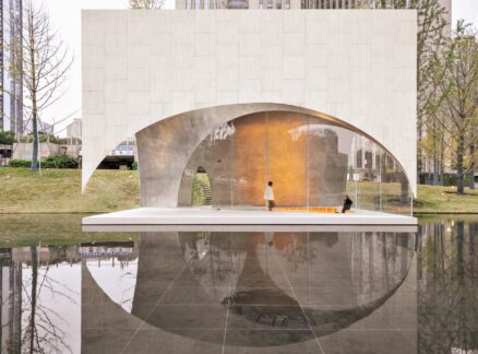 A modern structure with arched openings and textured walls stands by a reflective pool, with one person walking and another seated on an orange bench inside.