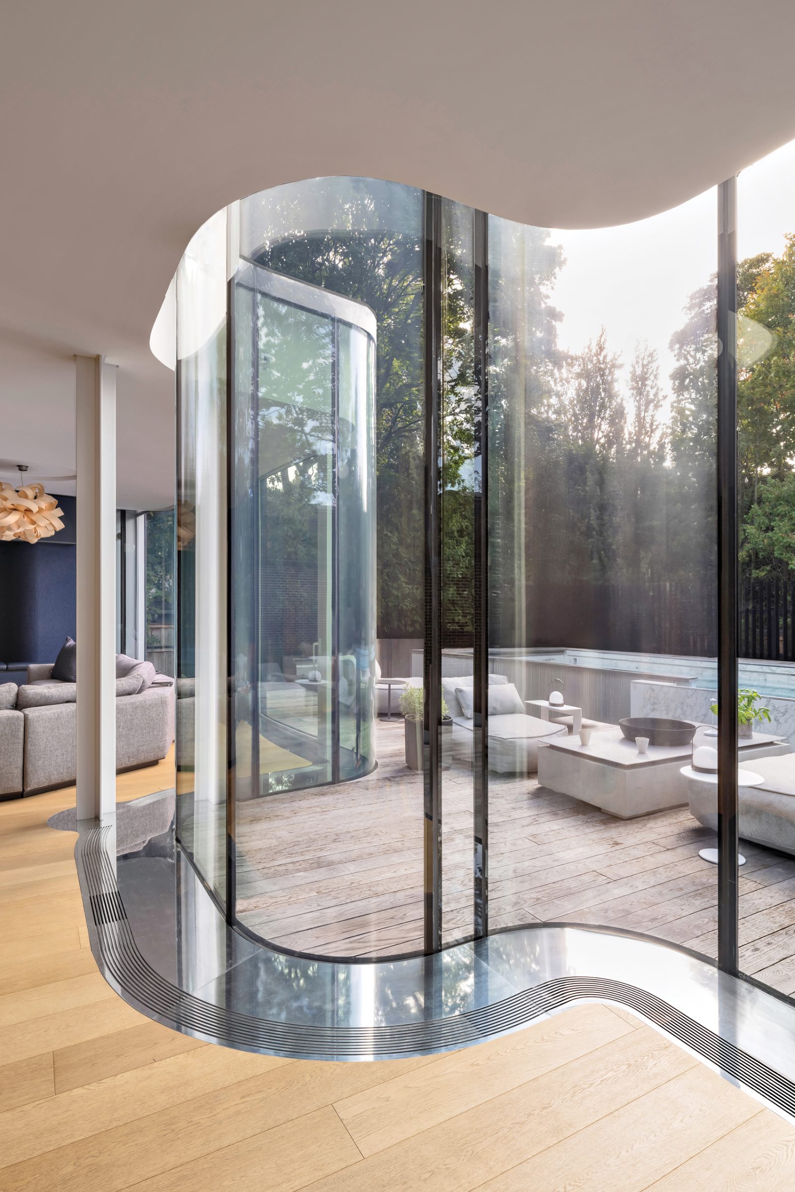 Curved floor-to-ceiling glass doors open to a wooden deck with outdoor seating; modern heritage home interior with light wood floors and neutral furniture visible.