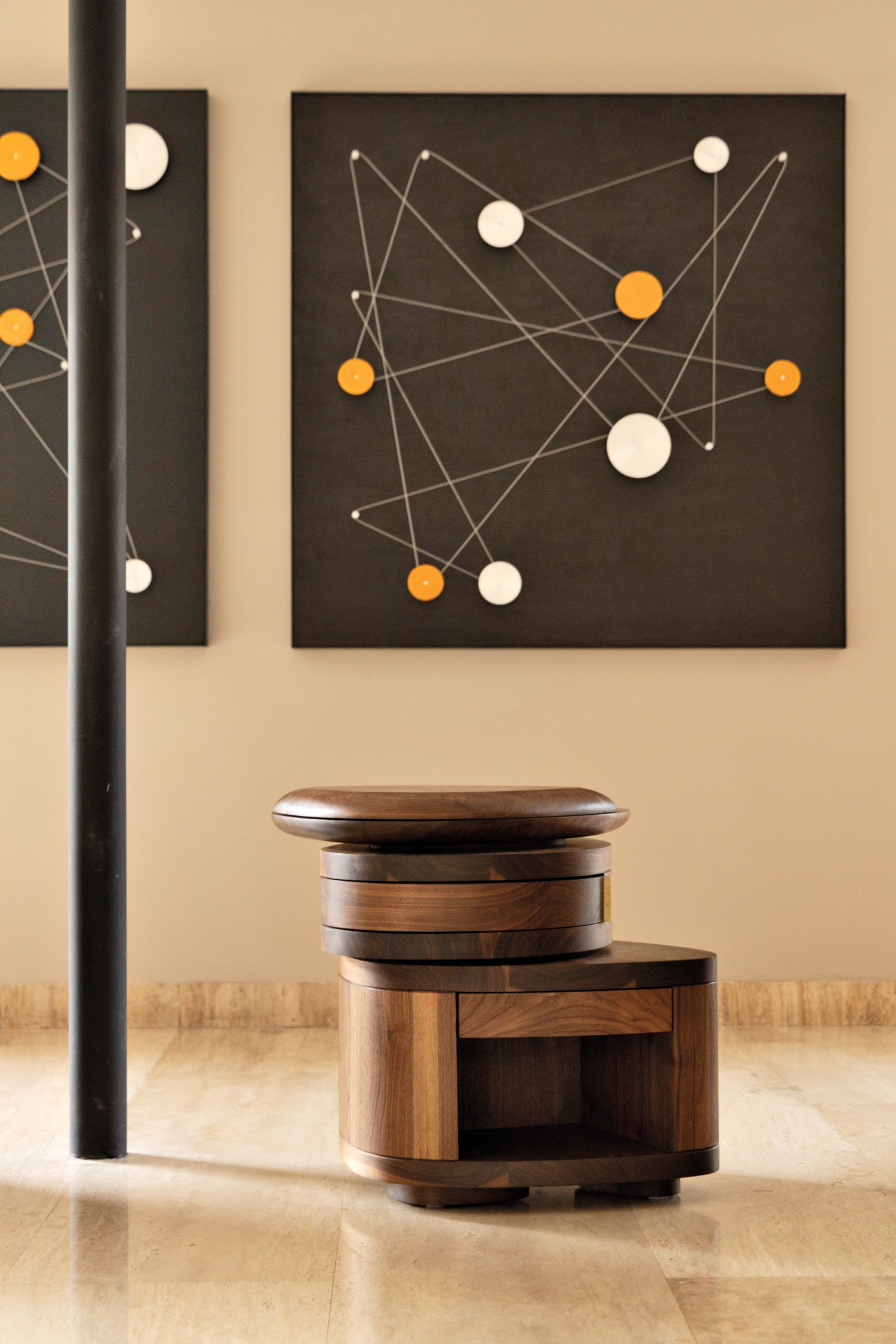 A modern wooden stool by Fernanda Canales sits on a beige floor in front of two geometric abstract artworks with white and orange circles connected by lines.