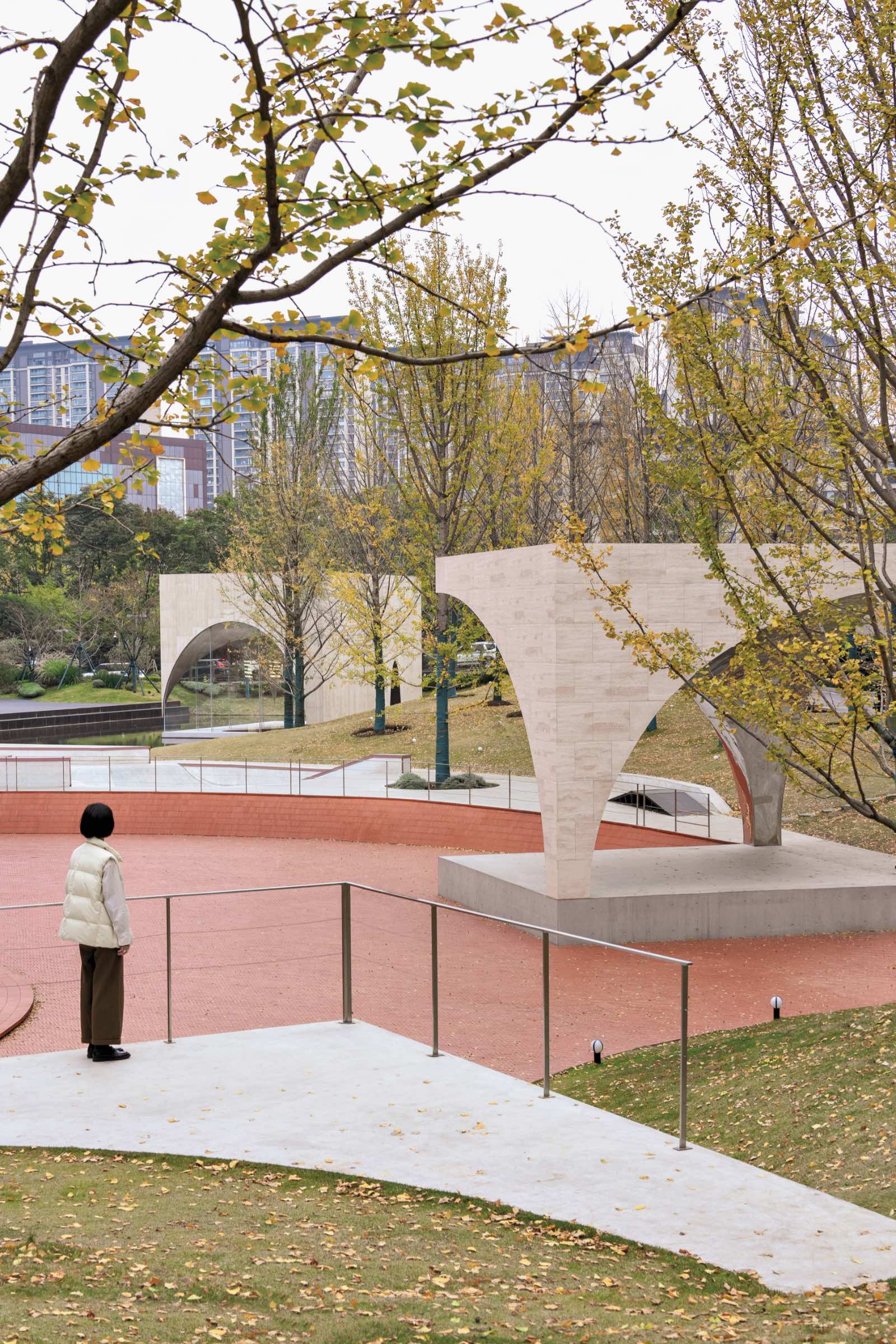 A person stands on a path near curved stone structures, AYDC Public Art Center by Atelier Xi, in a park with autumn trees and city buildings in the background.