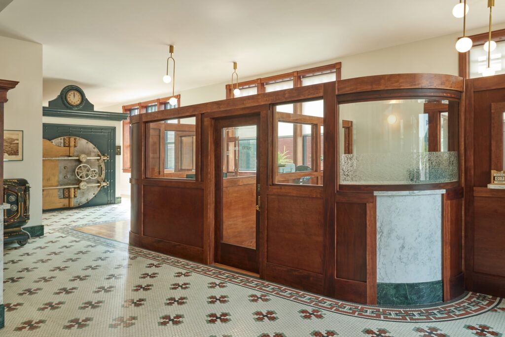 Historic bank interior with wood-paneled teller booths, mosaic tile floor, large vintage vault, and globe pendant lights.