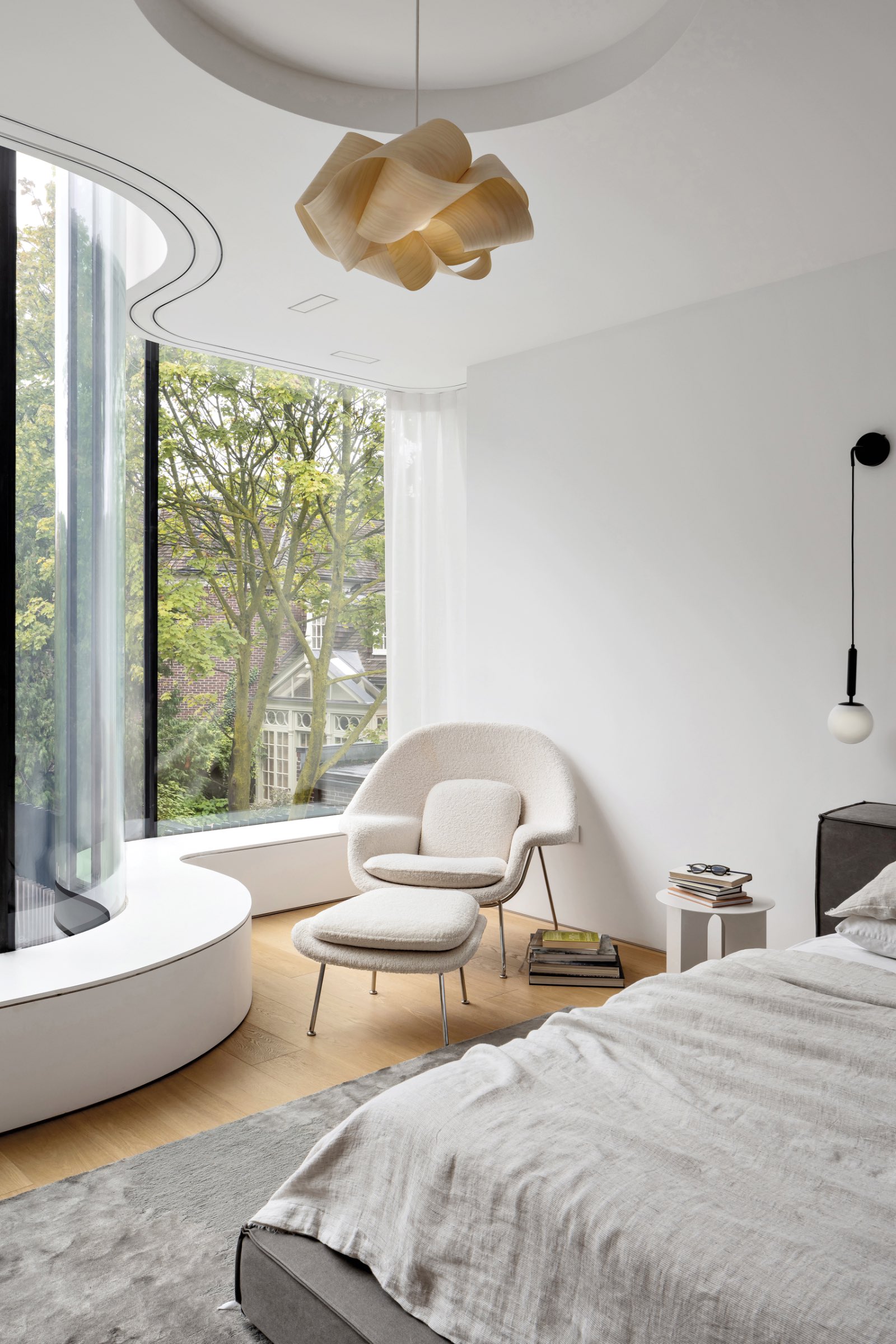 Minimalist bedroom in a heritage home with curved floor-to-ceiling window, modern lounge chair and ottoman, pendant light, small side table with books, and a view of trees outside.