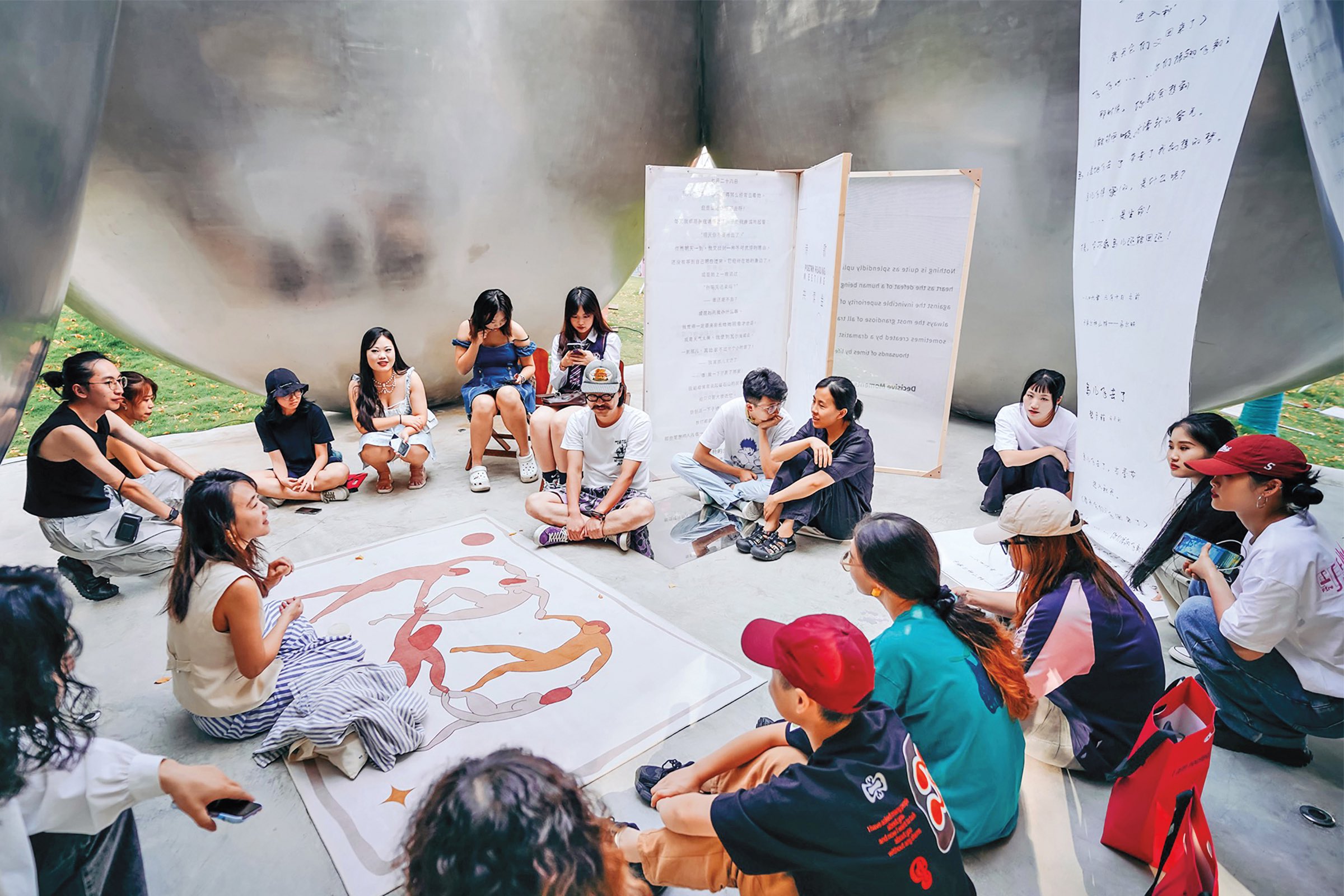A group of people sit in a circle on the ground, talking and listening, with posters and a large abstract artwork in the center.