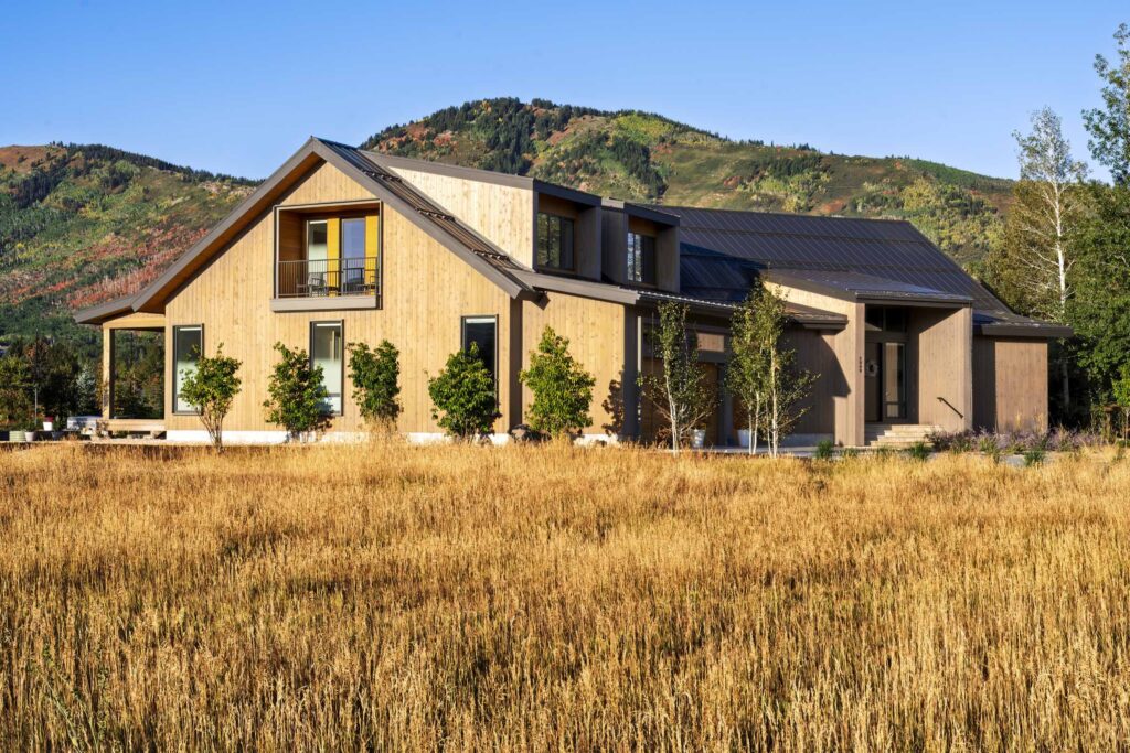Modern two-story house with large windows and sloped roof, surrounded by trees and tall grass, set against a backdrop of green hills and blue sky.