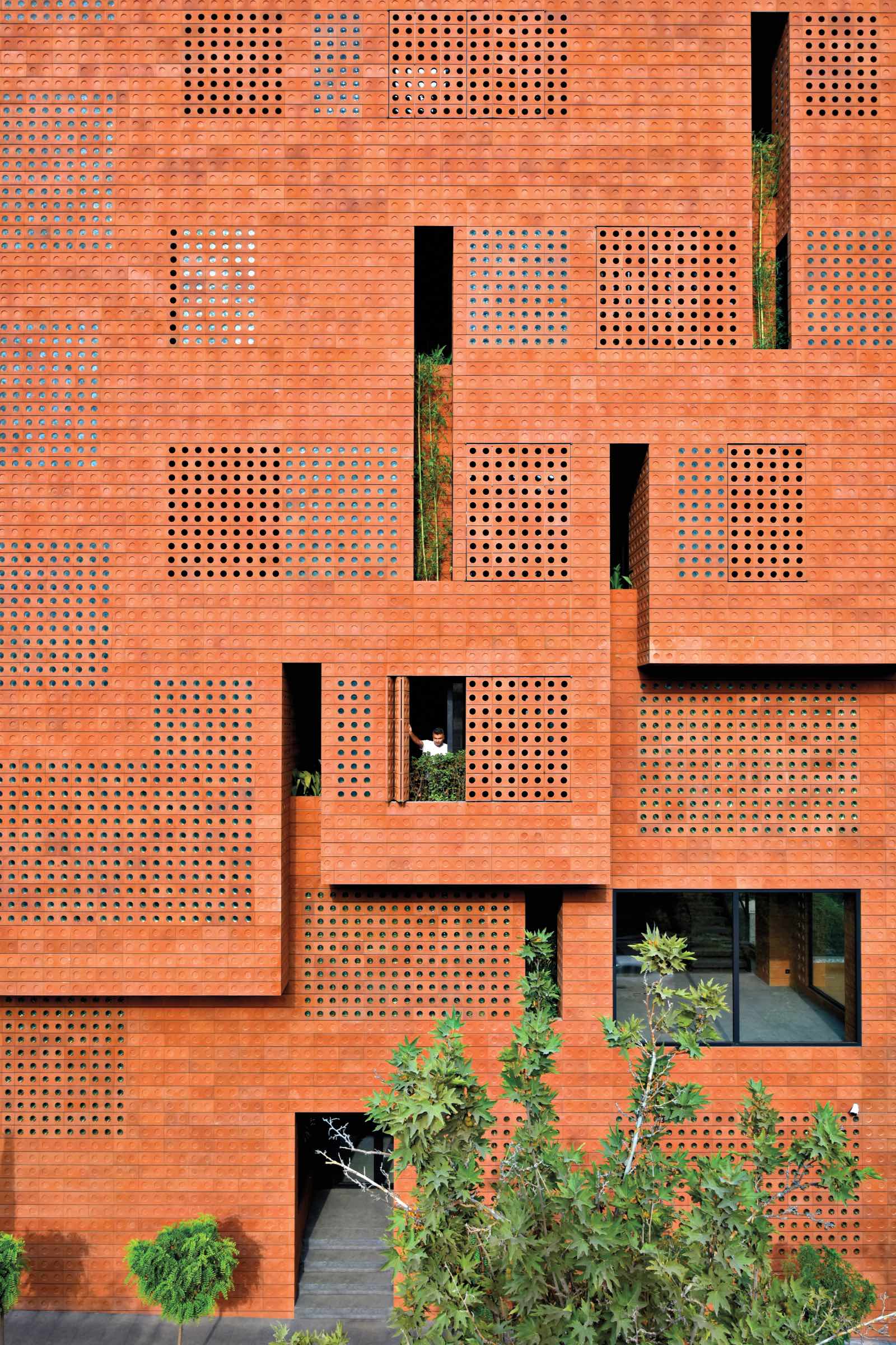 Red brick building facade by HOOBA Design Group with variously sized square-shaped perforations, some filled with plants, and a single window open with a person visible inside. Green foliage is seen at the bottom.