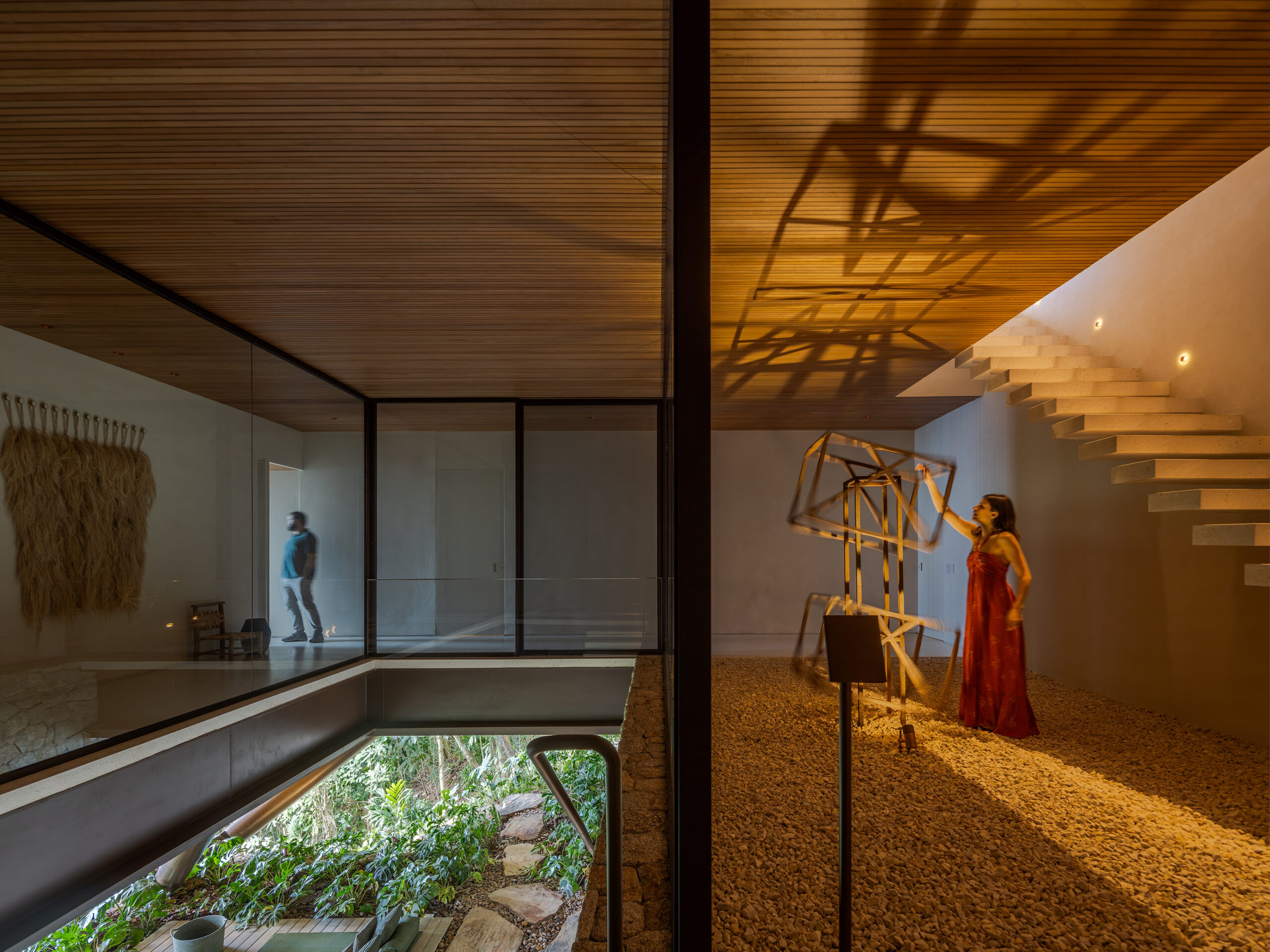 A woman in a red dress stands near a wooden sculpture under warm light; a man walks in a glass-walled hallway of this Passive House. Modern interior with stone, wood, and an indoor garden enhances the sustainable elegance of the space.