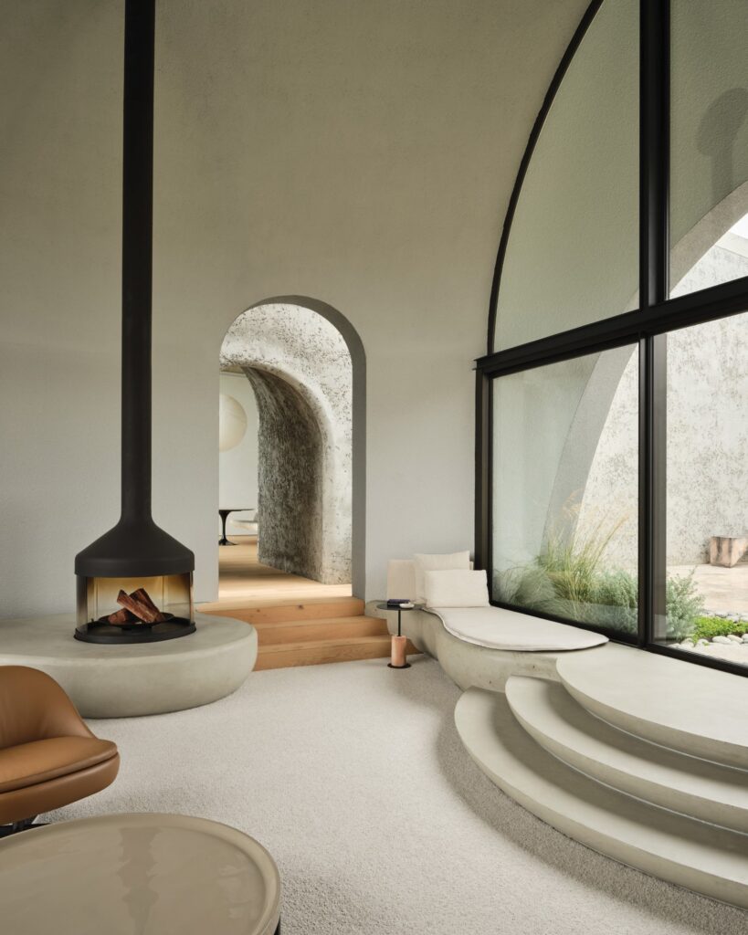 Modern living room with minimalist furniture, a suspended fireplace, curved stairs, large arched window, and textured concrete walls. Natural light fills the space.