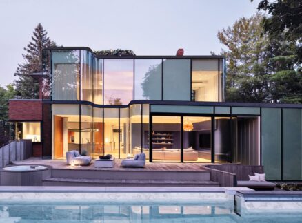 Modern two-story heritage home with large glass windows, outdoor seating, and a pool in the foreground, surrounded by trees.