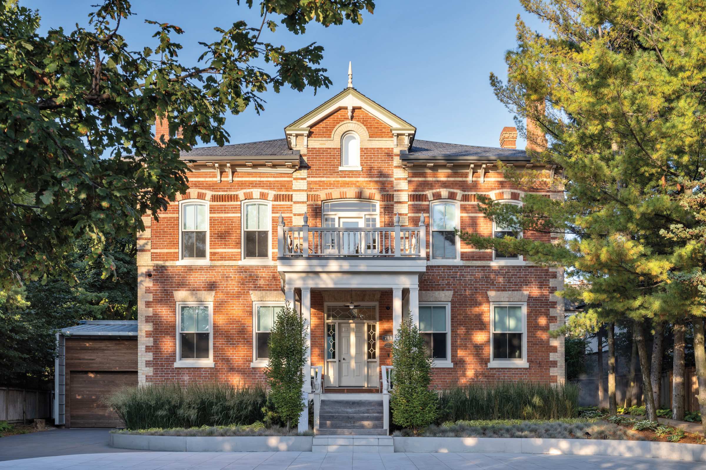 Two-story red brick heritage home with white trim, a central porch, balcony, symmetrical windows, and a garage on the left, surrounded by trees and landscaping.